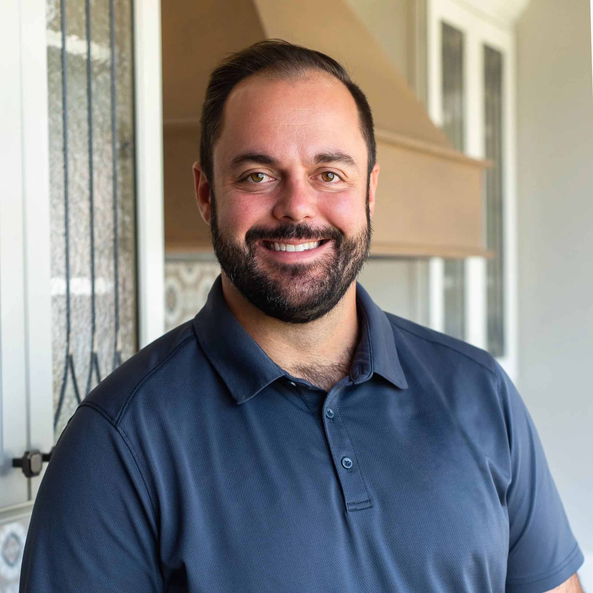 Close-up of a smiling man with a beard, wearing a dark blue button-up shirt, standing in front of a house with a window and a porch.