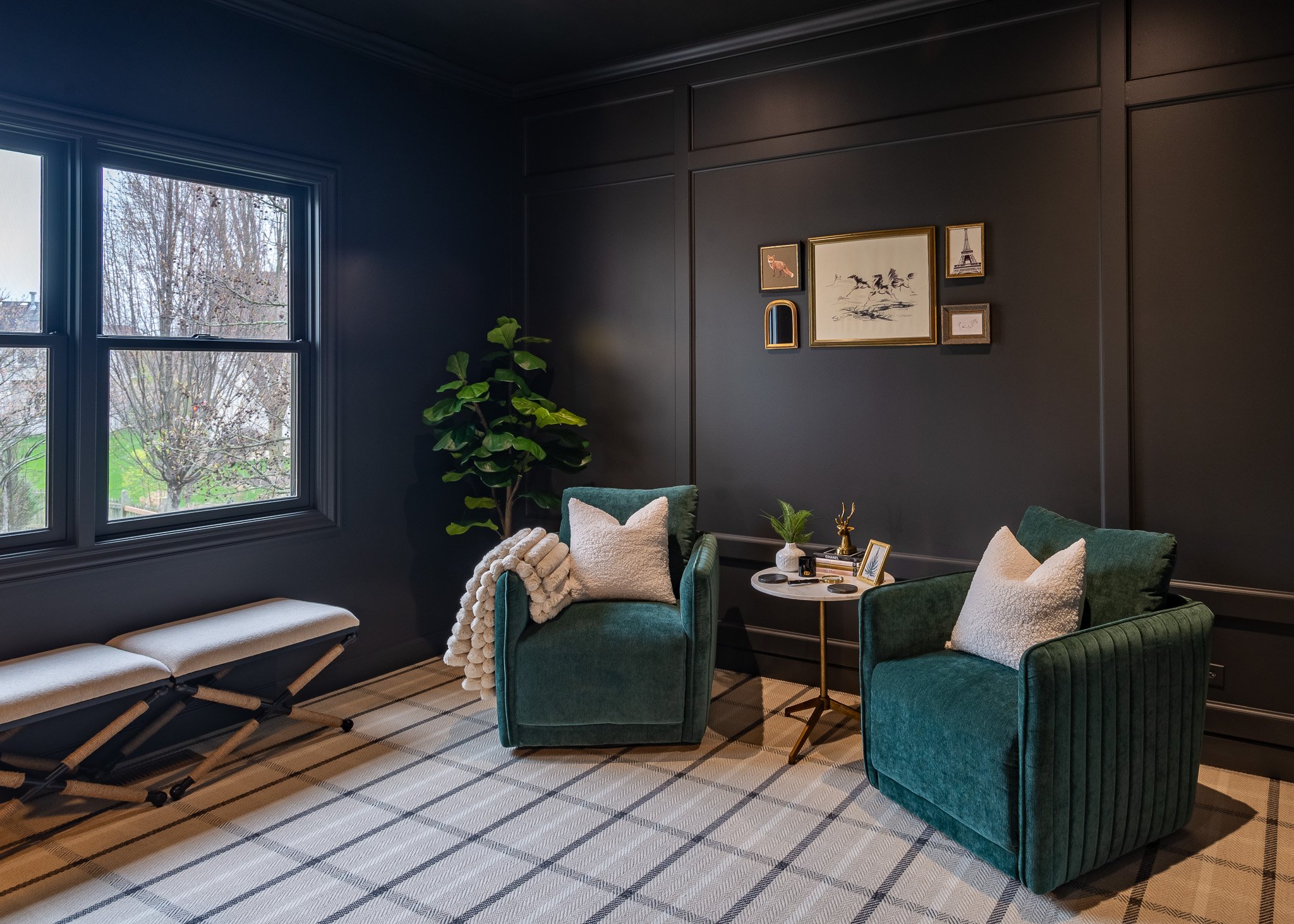 Cozy living room with dark walls, green velvet armchairs, a beige upholstered bench, framed artwork including the Eiffel Tower, a potted plant, and a window showing bare trees outside.