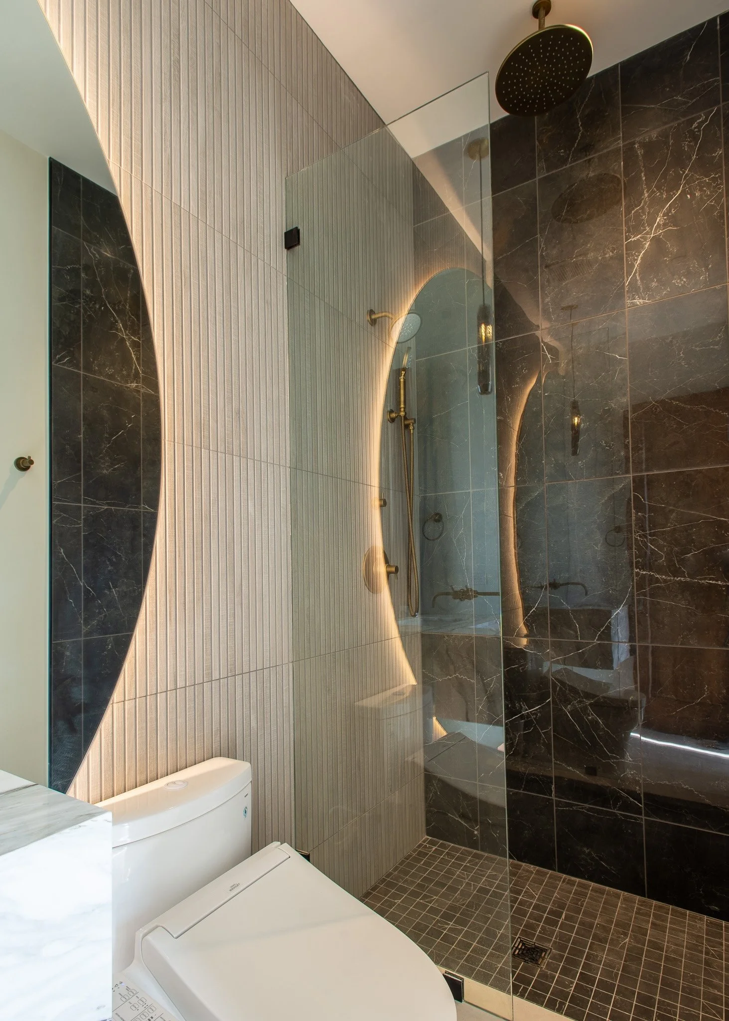 A modern bathroom with a shower area separated by a glass partition, a black marble wall, a beige tiled wall, and a white toilet. The shower has a large black rain showerhead and brass fixtures.