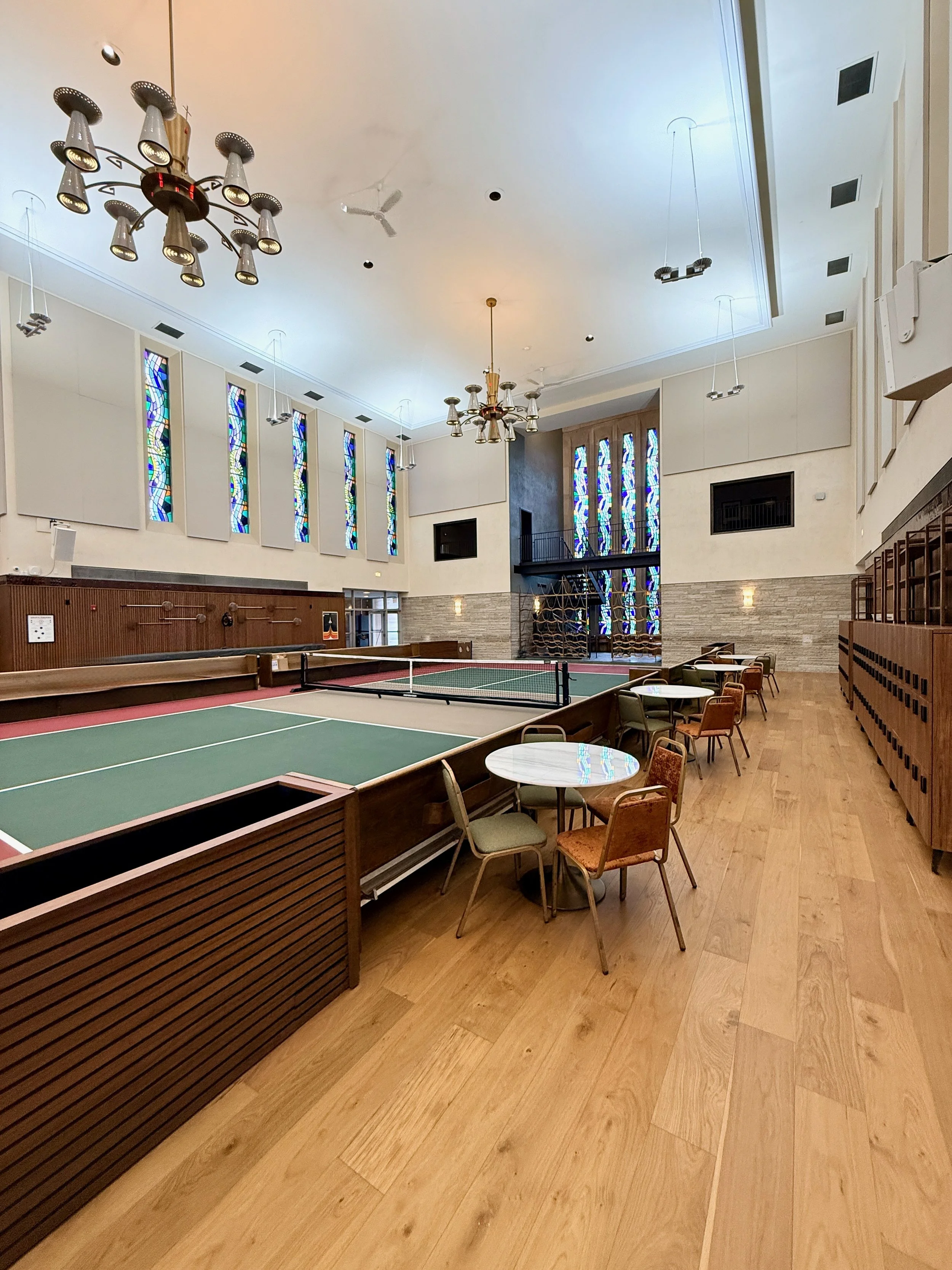 An indoor recreational space with a ping pong table, chairs, tables, large windows with colorful stained glass, high ceilings, and chandeliers.