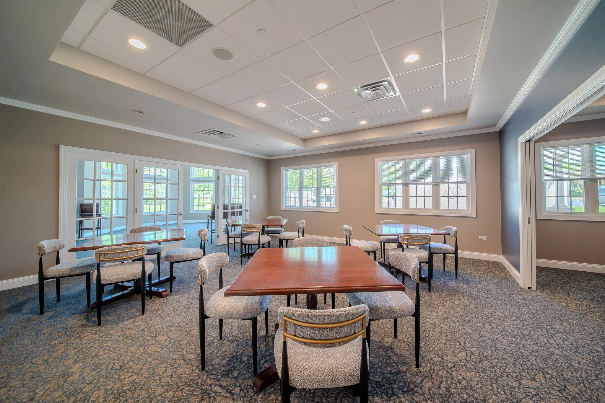 Meeting or conference room with multiple chairs arranged around tables, large windows with blinds, and a ceiling with recessed lighting and air vents.