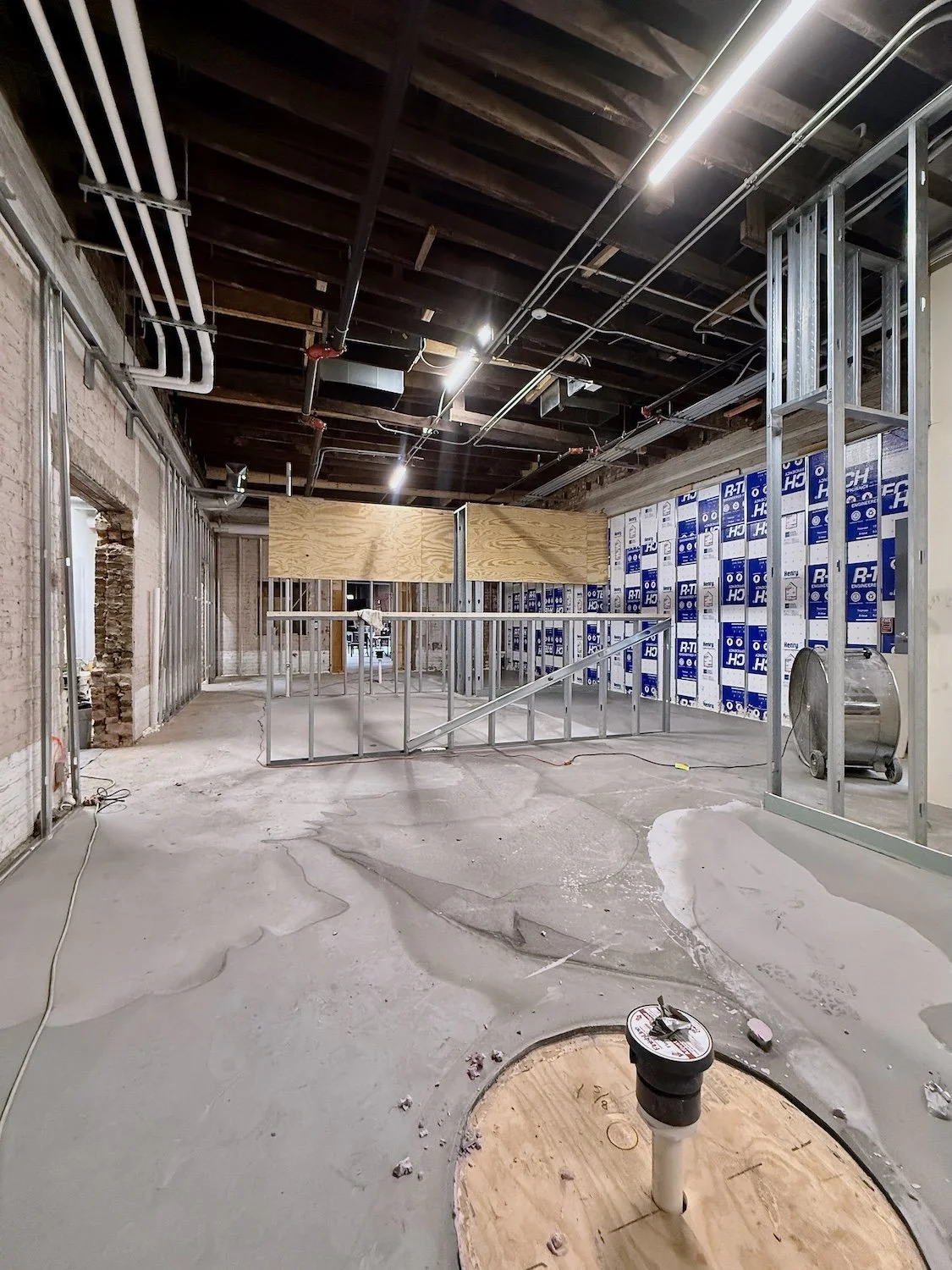 Interior of a building under construction with exposed framing, pipes on the ceiling, and unfinished floors.