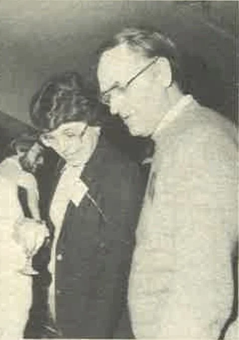 Two individuals, a woman and a man, wearing glasses and face masks, looking down and smiling, likely engaged in a conversation or an activity.