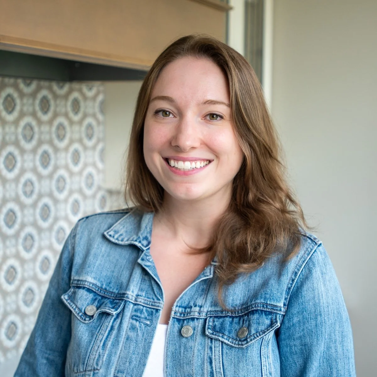 A woman with shoulder-length brown hair wearing a denim jacket, smiling at the camera, indoors with patterned wallpaper in the background.