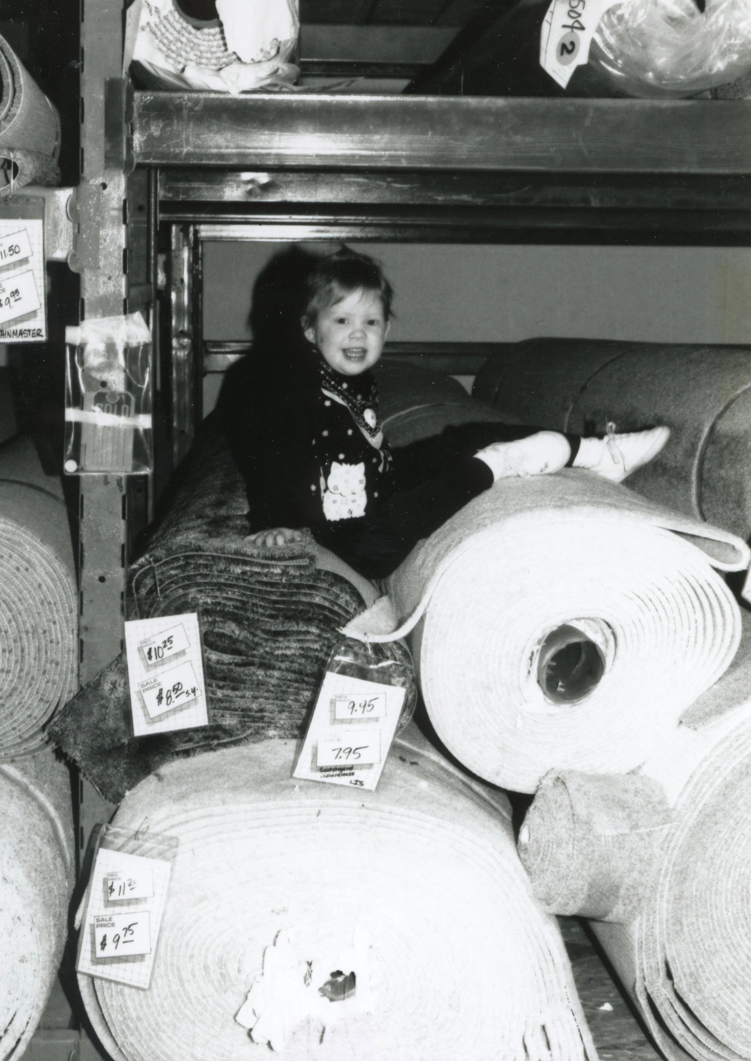 A young girl with short hair and a big smile sitting on a rolled rug in a store with various rugs and price tags around her.