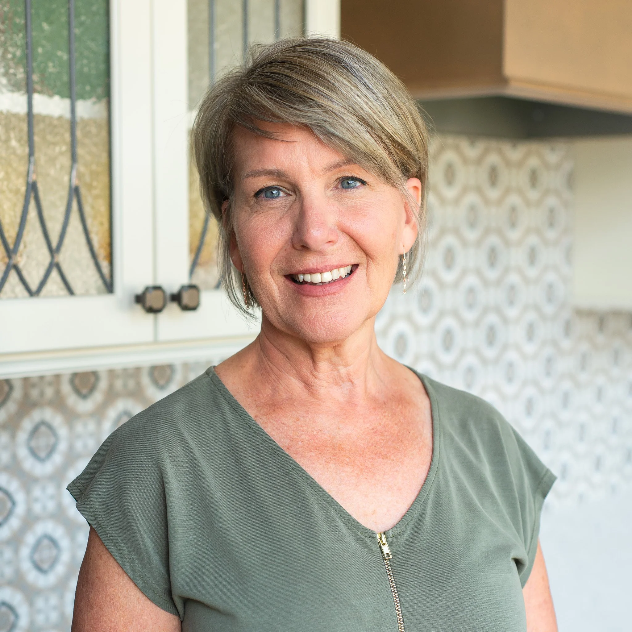 Close-up of a smiling middle-aged woman with short blonde hair, wearing a green top with a zipper, standing in front of a window with a decorative wall pattern.