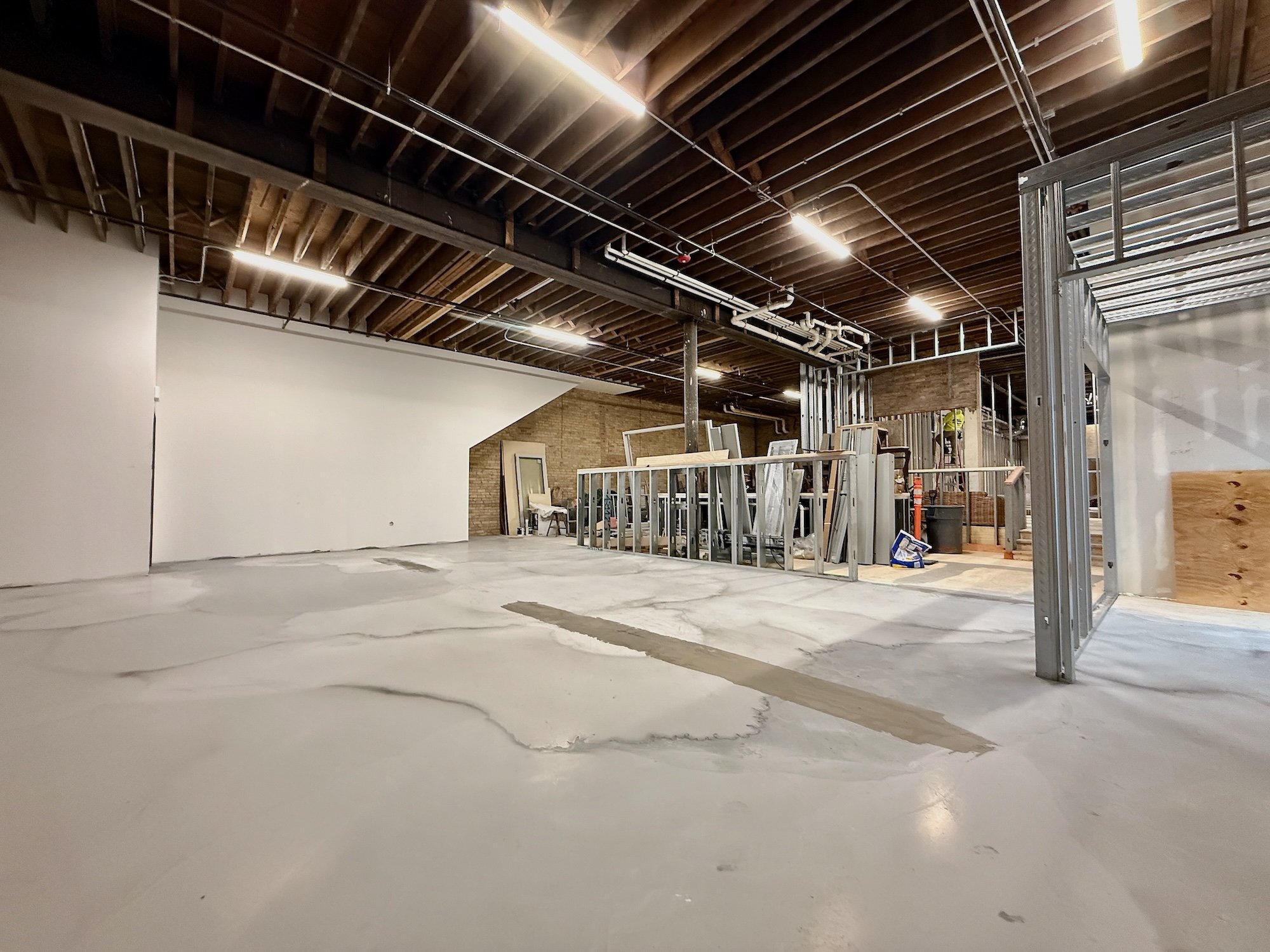 Interior of a building under construction with visible ceiling pipes, framing, and unfinished concrete floors.