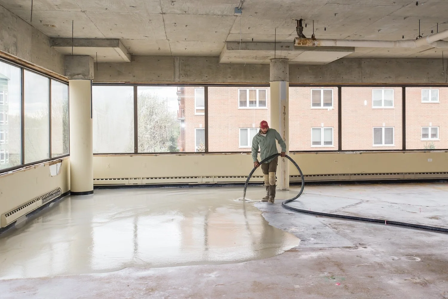 A man in casual work clothes and a red cap is pouring a liquid substance on a concrete floor with a large hose inside a room with large windows and an unfinished ceiling.