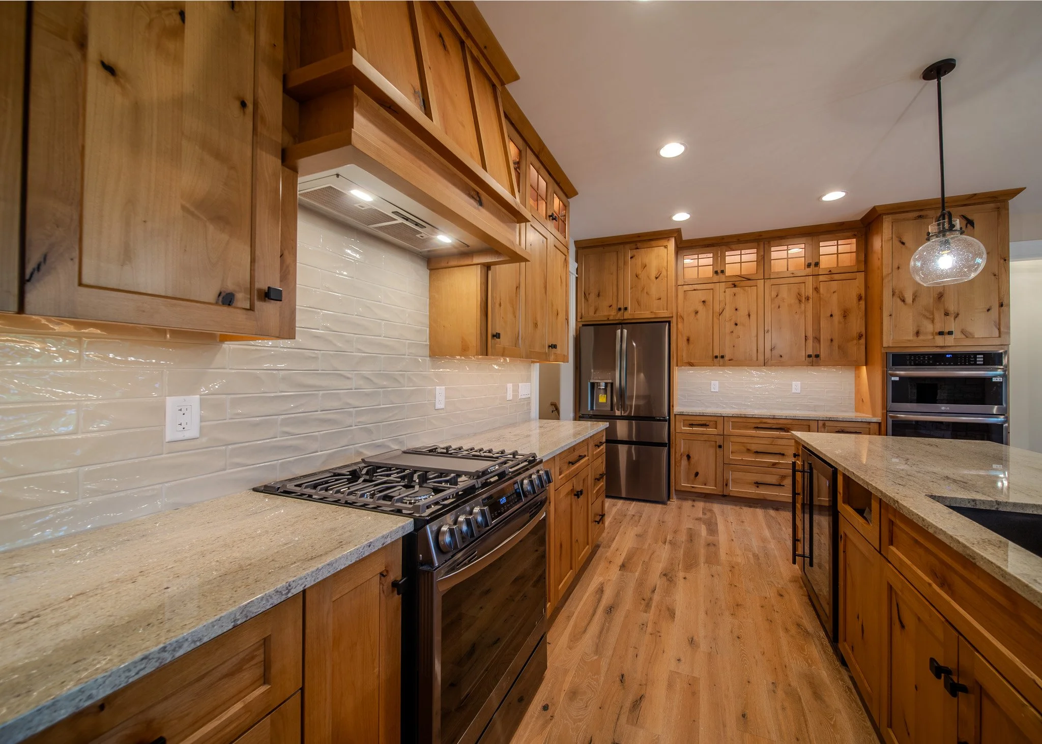 A spacious kitchen with wooden cabinets, beige countertops, a stainless steel refrigerator, a gas stove, and modern lighting fixtures.
