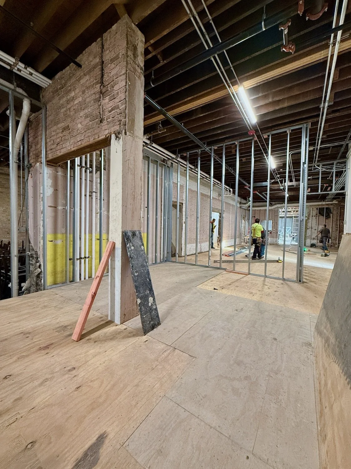 Construction site with exposed brick and metal framing for walls, workers installing drywall, and construction materials on the floor.