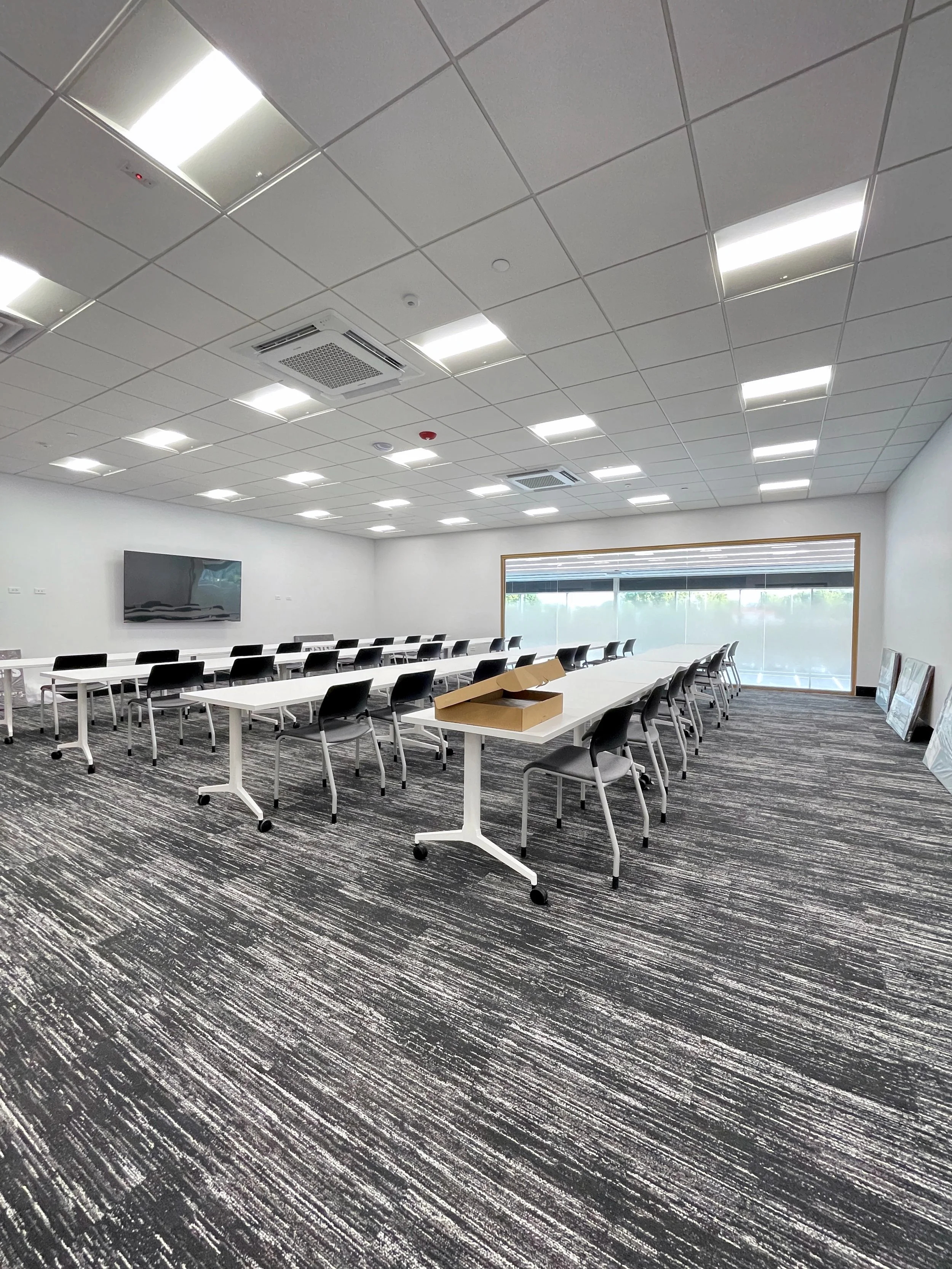 A modern conference room with white tables, black chairs, a flat screen TV, and a window with blinds.