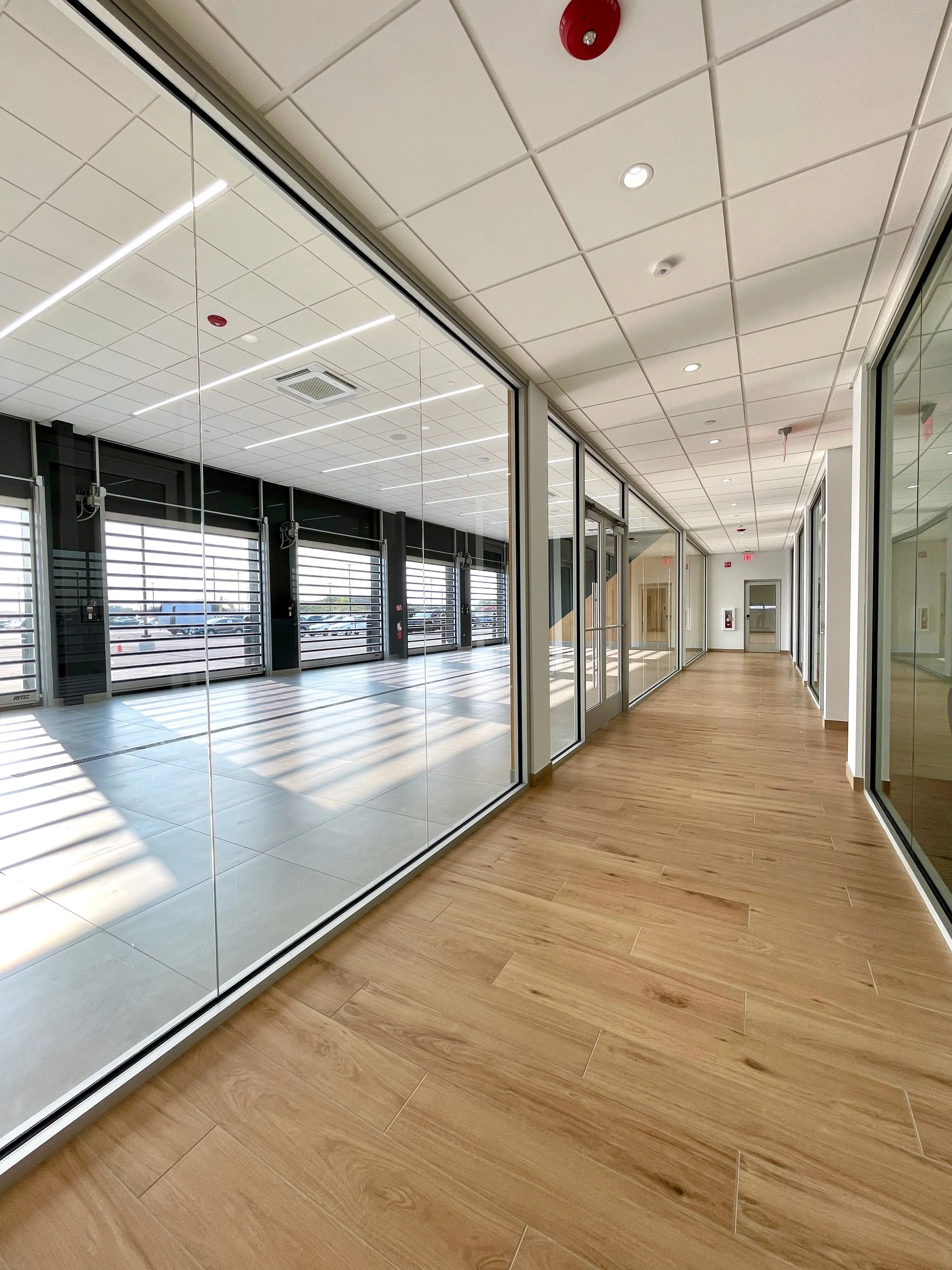 A hallway in a commercial building with large glass windows and doors, wooden flooring, and a ceiling with recessed lighting.