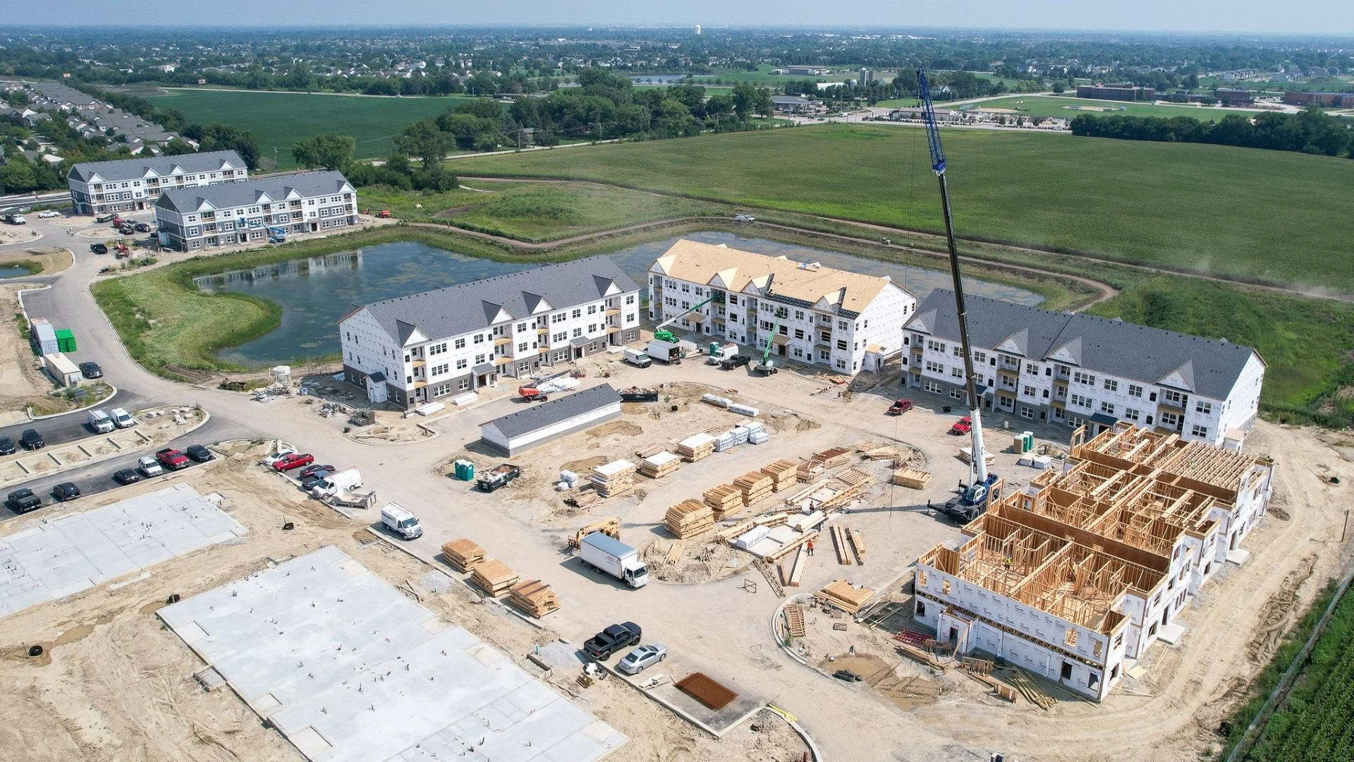 Aerial view of a construction site with multiple multi-story buildings under development, surrounded by construction equipment, materials, and parked vehicles, with green fields and residential areas in the background.