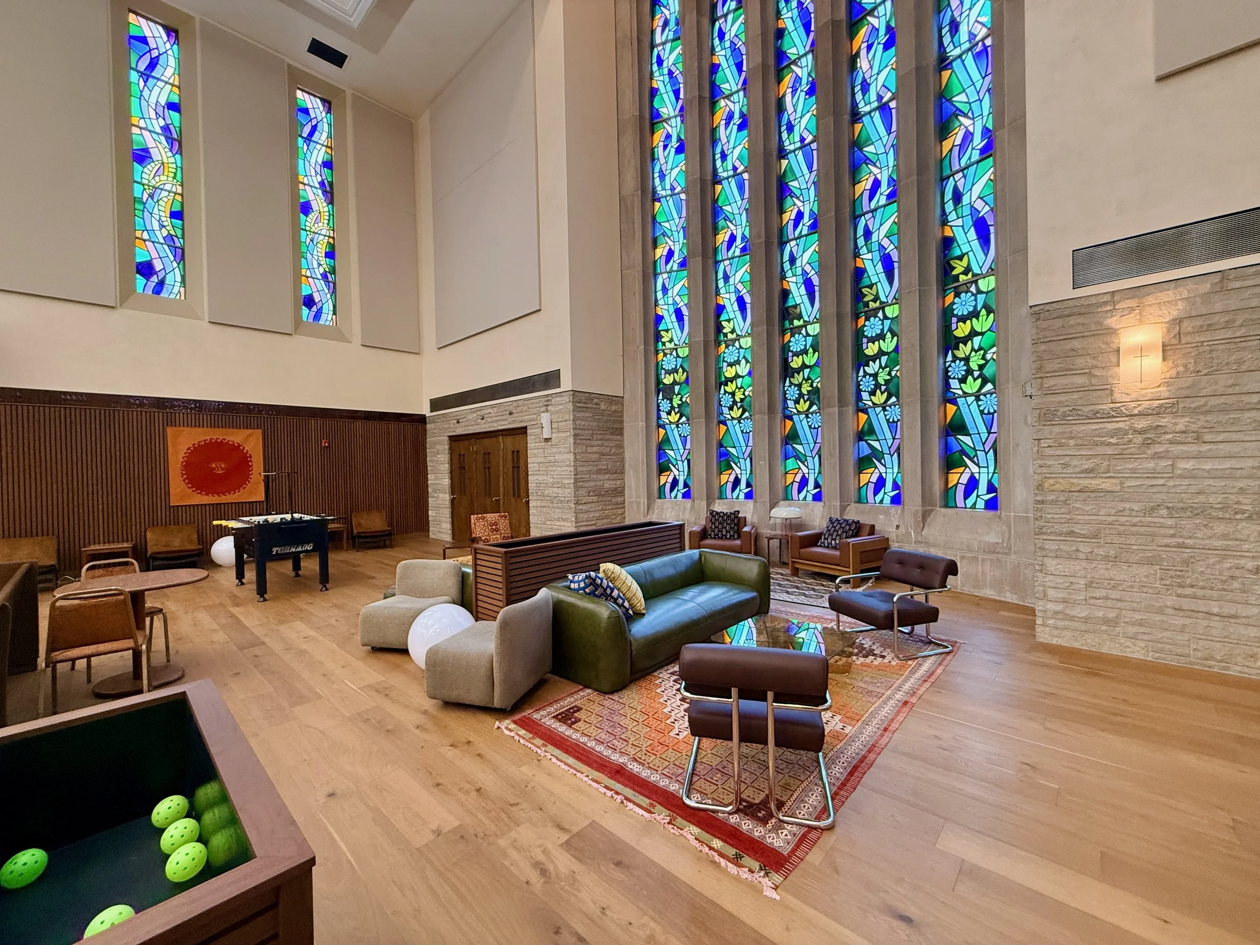 Interior of a church or chapel with large colorful stained glass windows depicting abstract and floral designs, a cozy seating area with modern furniture, a rug, and a foosball table with green balls in the foreground.
