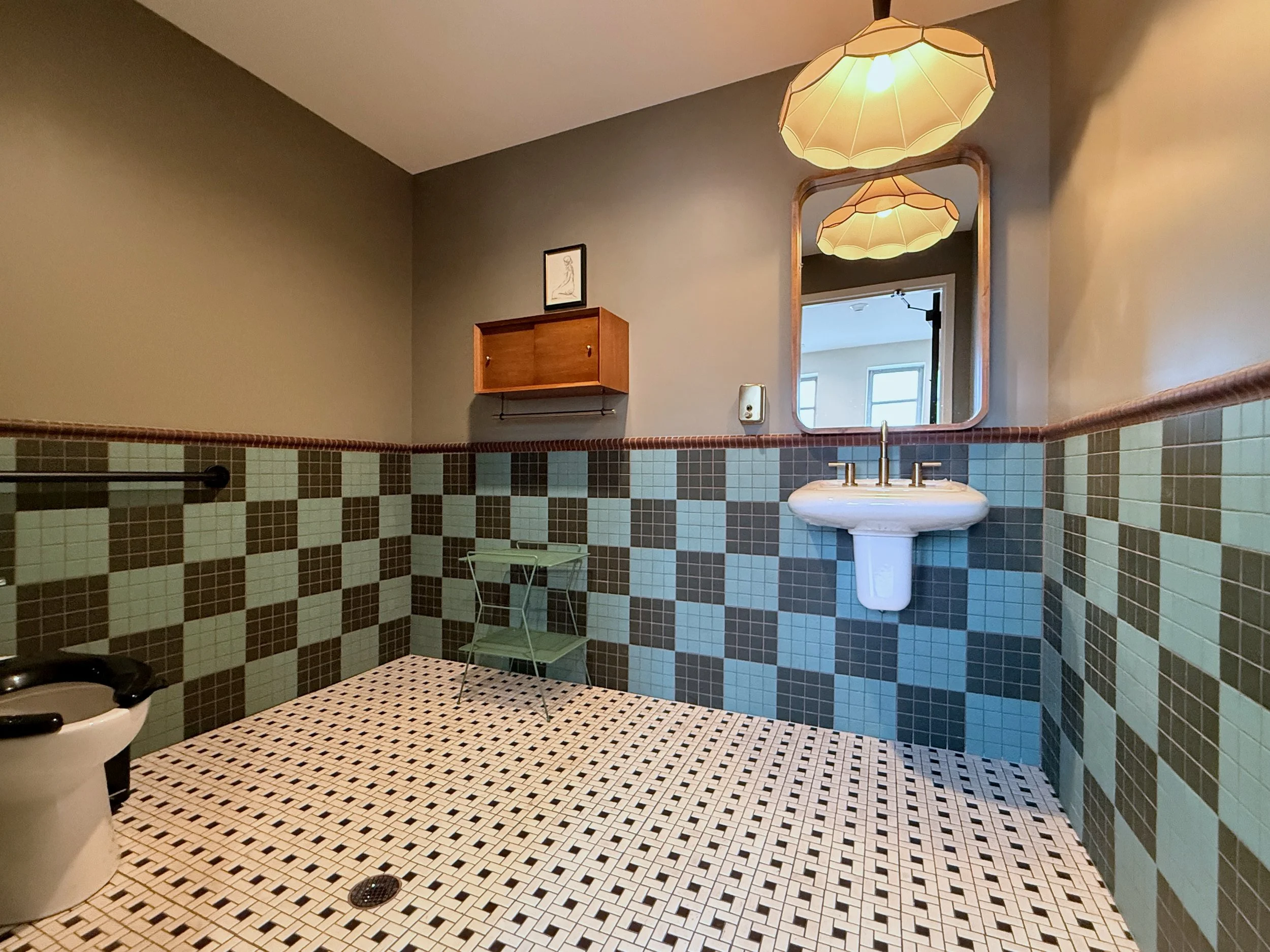 Bathroom with green and brown tiled walls, a white pedestal sink, a mirror, a wooden wall cabinet, a hanging light fixture, and a mirrored door.
