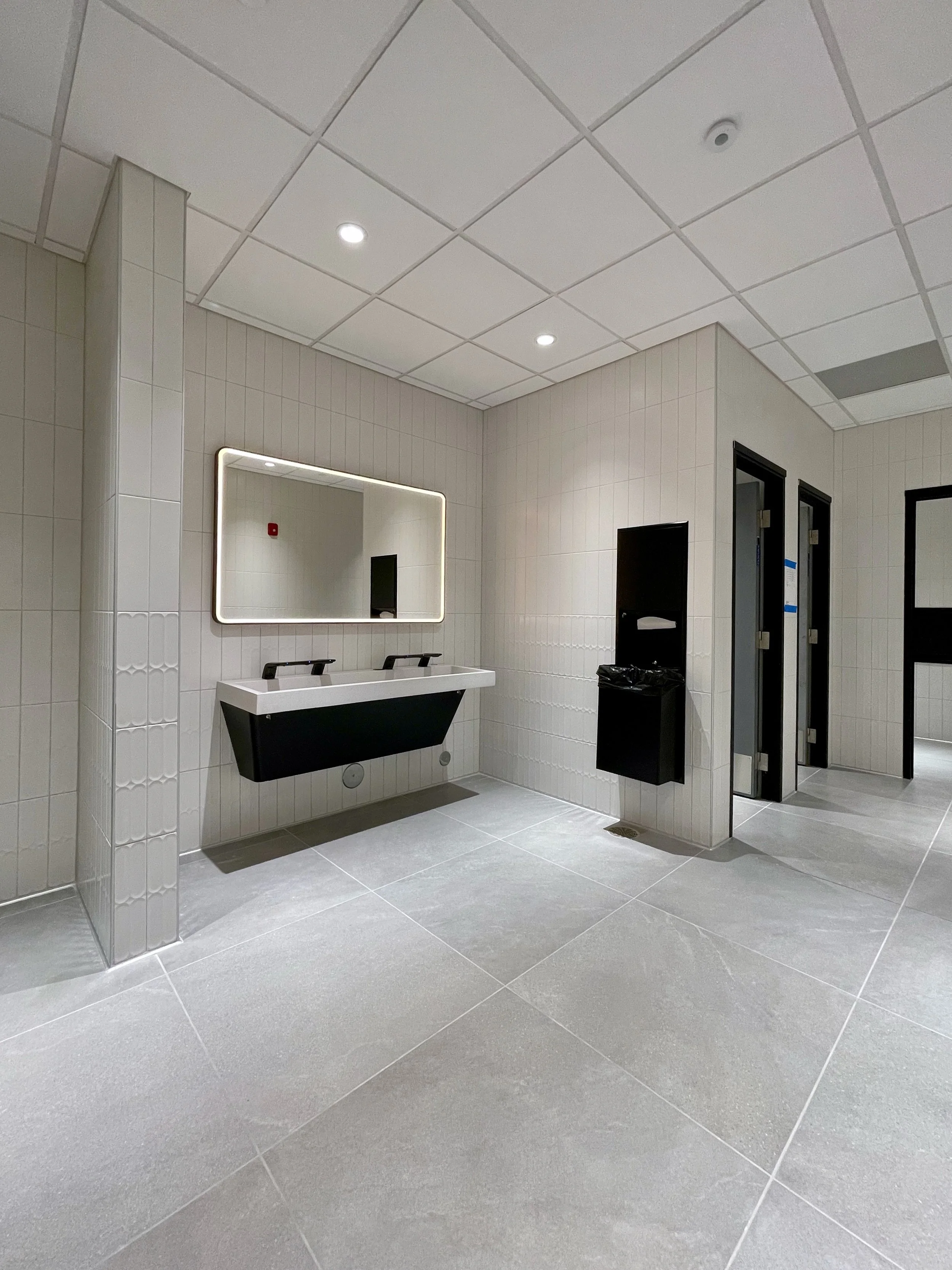 Public men's restroom with a white sink, large mirror with lighting, and two toilet stall doors, tiled walls and floor.