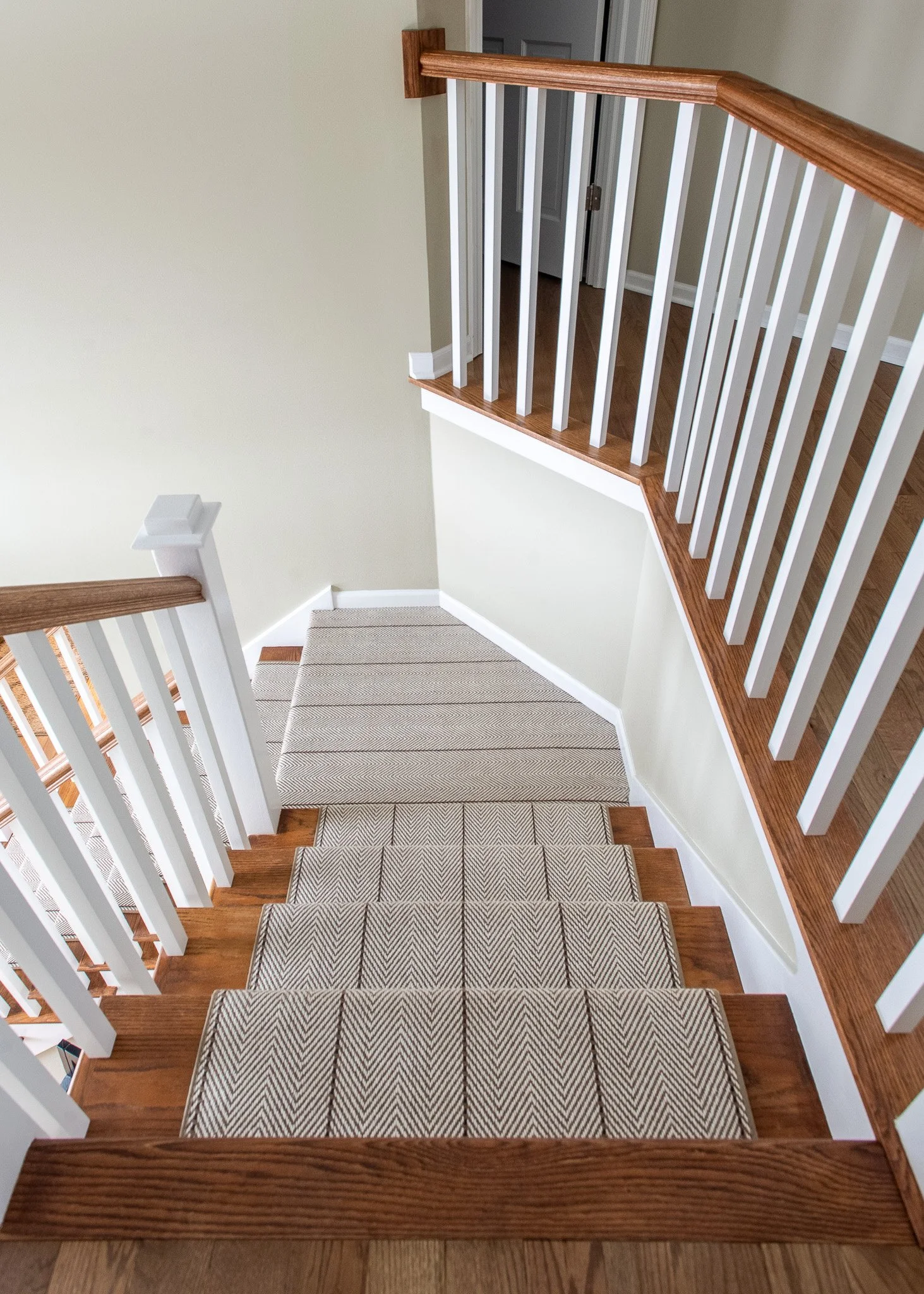 View of a staircase with wooden handrails and beige carpeting, leading down to a door at the bottom of the stairs, with an off-white wall and hardwood flooring.