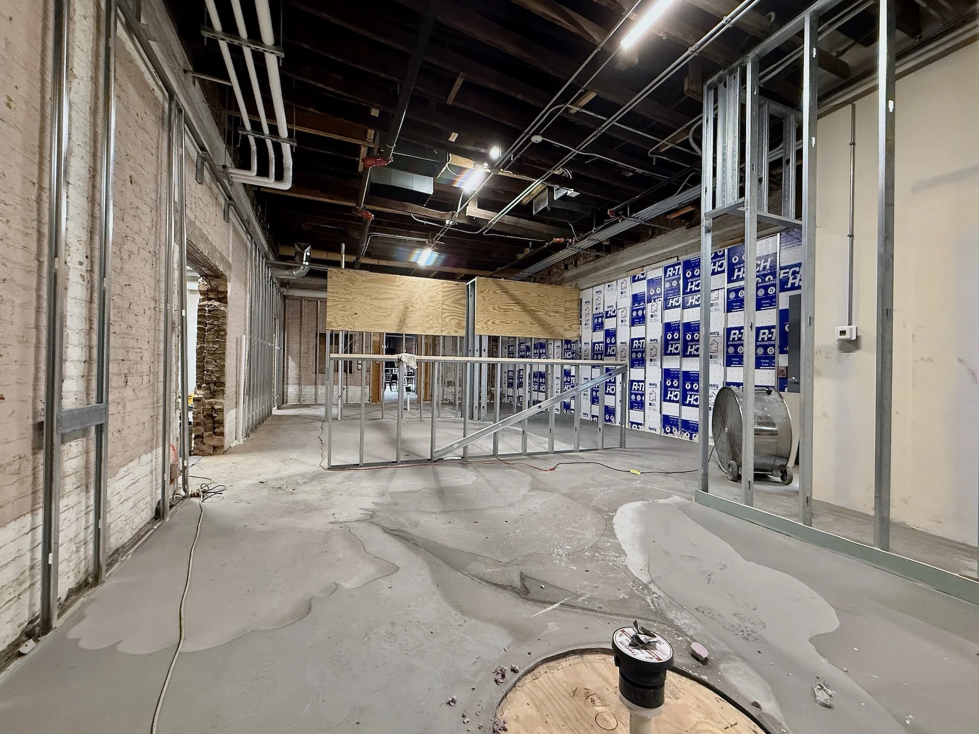 Construction site with exposed brick walls, metal framing, and a partially finished ceiling. There are pipes, electrical conduits, and insulation visible, with some areas covered with blue and white material and a wooden partition in the background.