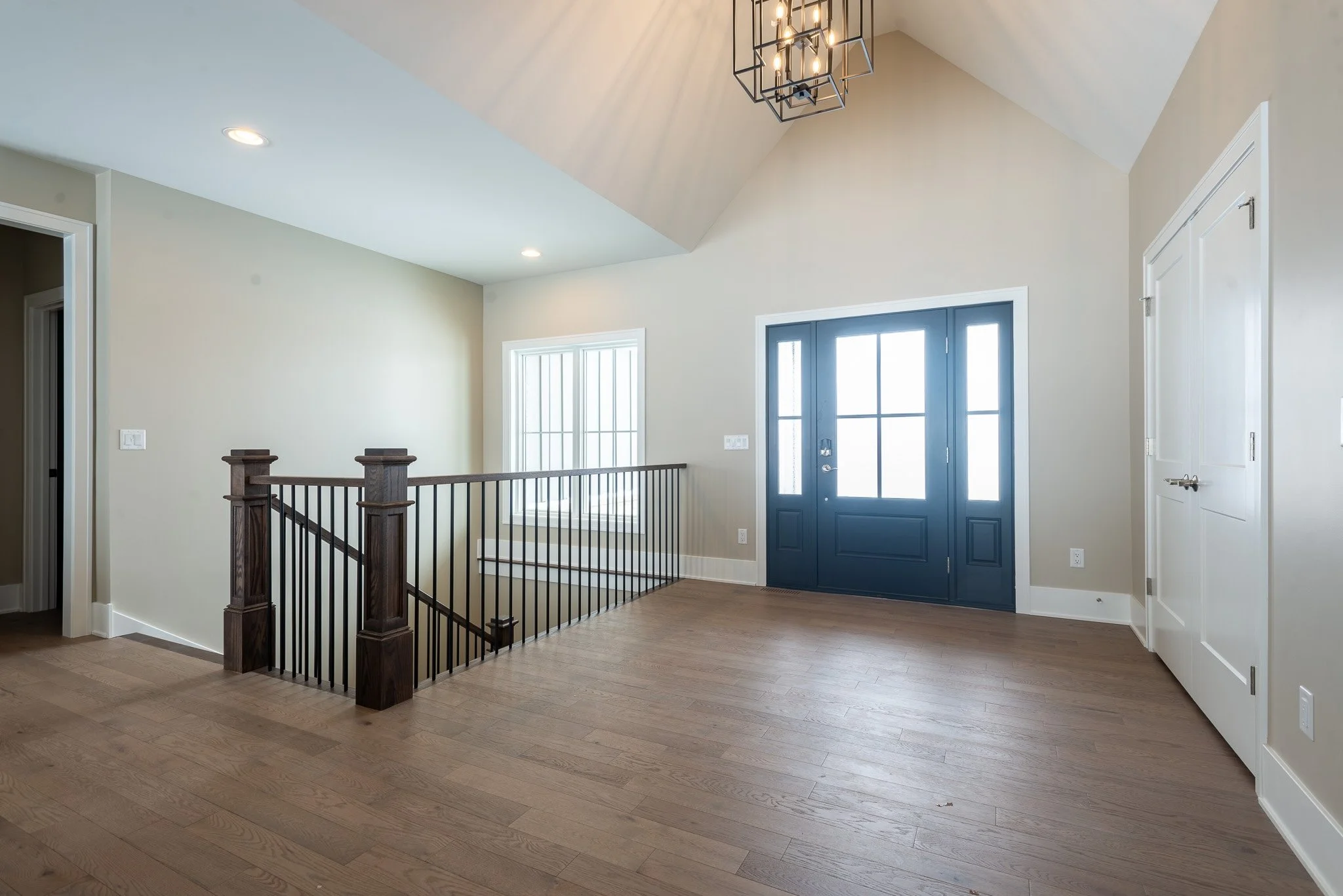 Interior view of a modern house entryway with a front door, a window, hardwood floors, and a staircase with dark wooden posts and black metal railing.