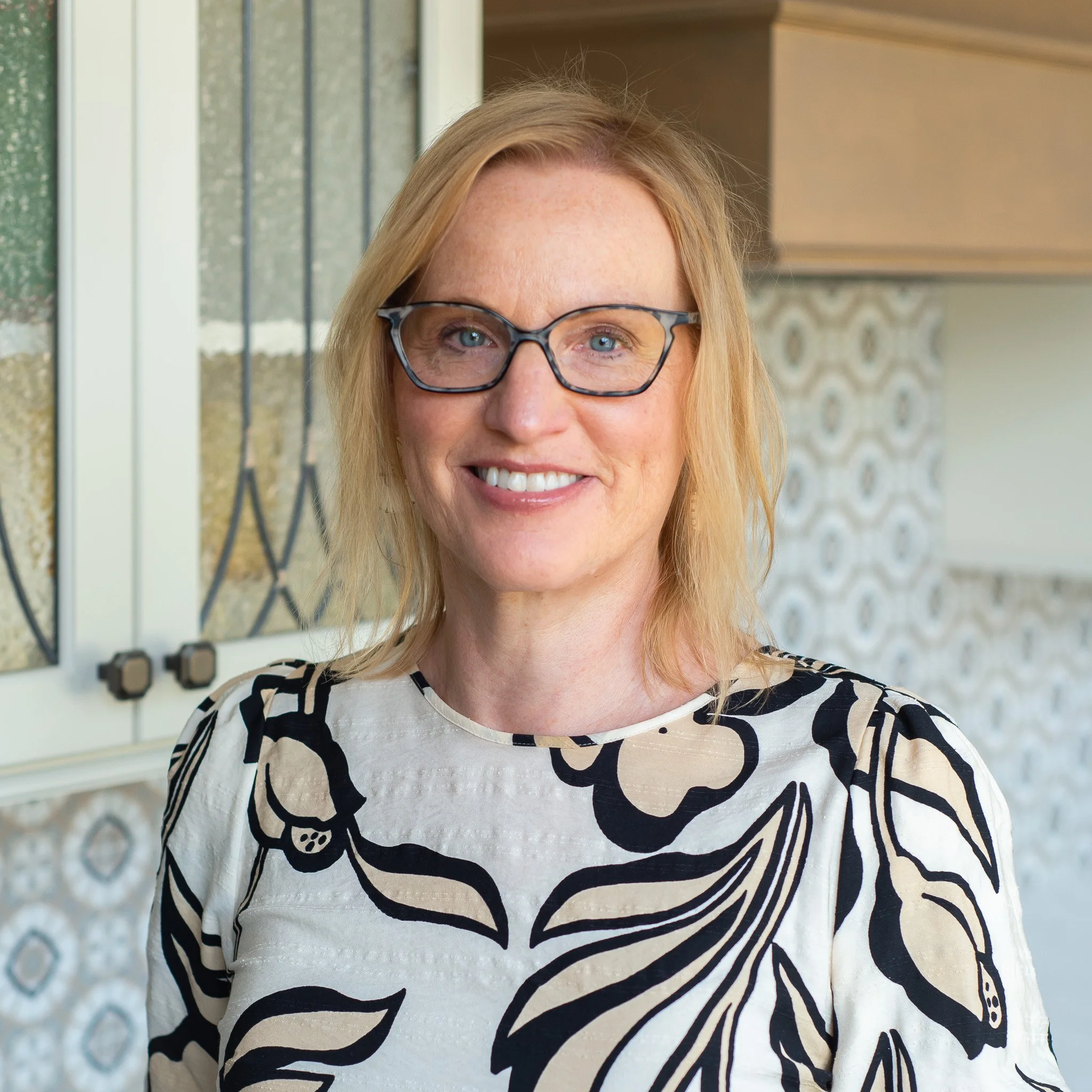 A woman with blonde hair and glasses smiling outside, wearing a black and white patterned top.