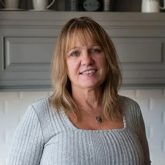 A woman with shoulder-length blonde hair, wearing a gray ribbed sweater and a necklace, standing in front of a gray mantel with decorative objects.