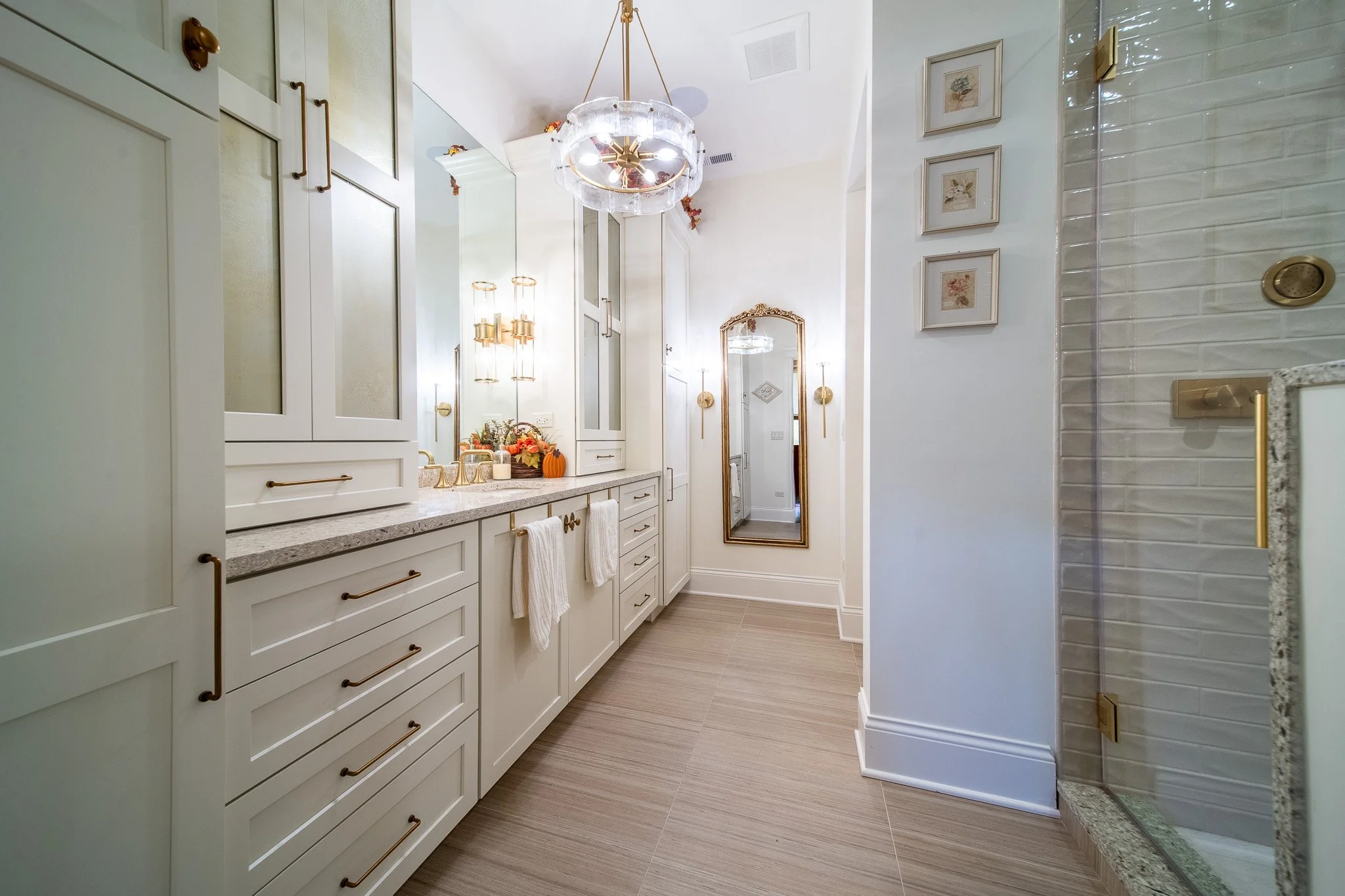 A bright bathroom with white cabinets, a granite countertop, gold fixtures, a large mirror, and wall-mounted lights. Decor includes small framed art, a chandelier, and fall-themed decorations such as a pumpkin and flowers near the sink.