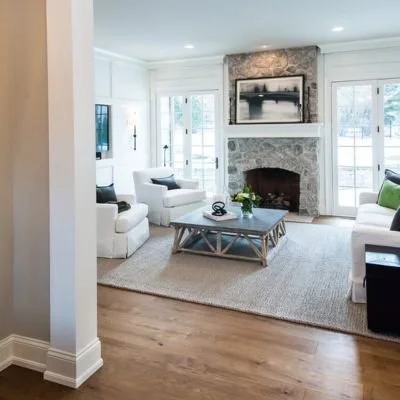 Living room with white sofas, a stone fireplace, a coffee table, a large painting above the fireplace, and windows with white shutters.