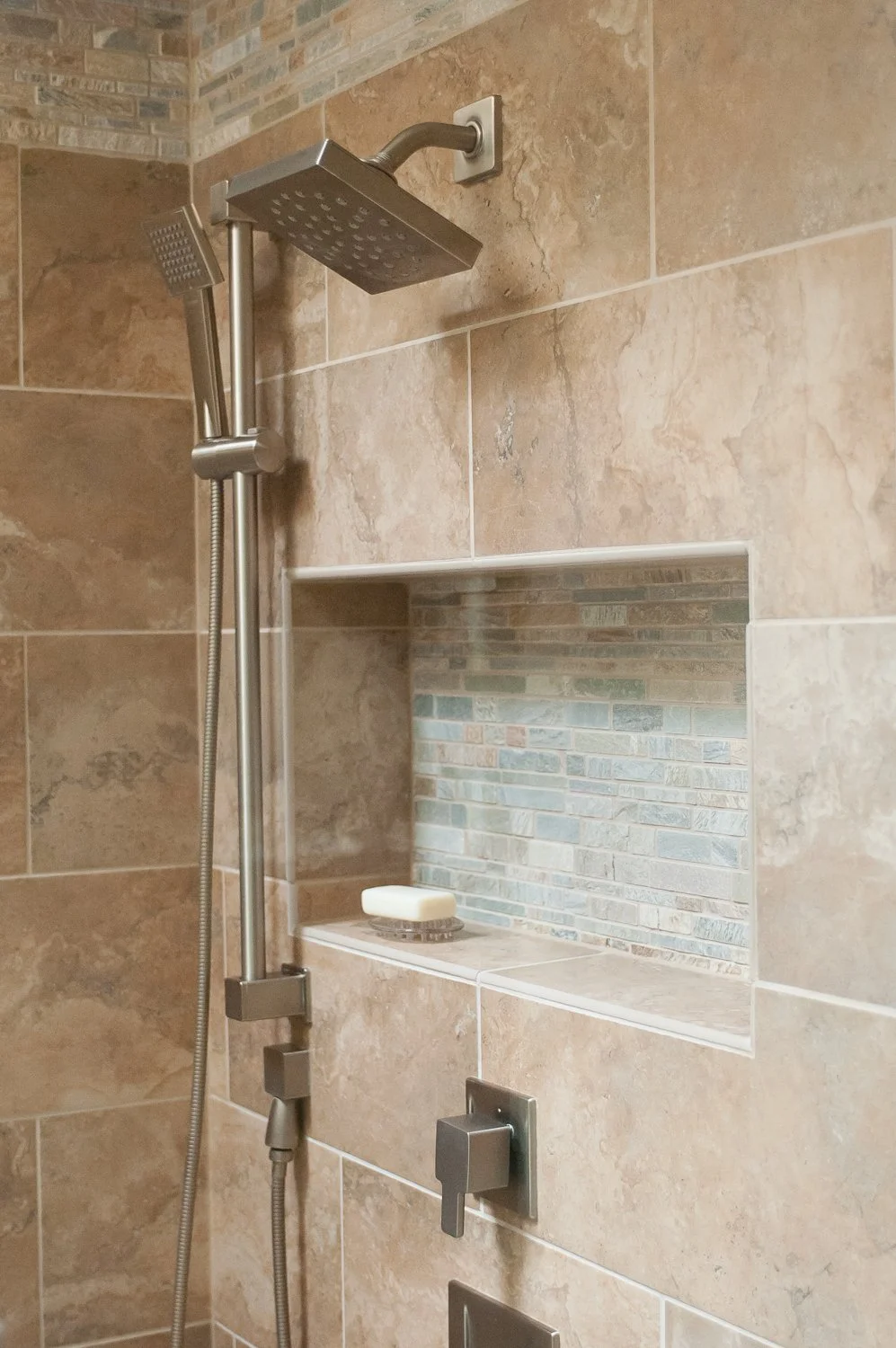 A shower with dual showerheads and a built-in shelf with a bar of soap, set in a tiled bathroom wall with beige and brick pattern mosaic tiles.