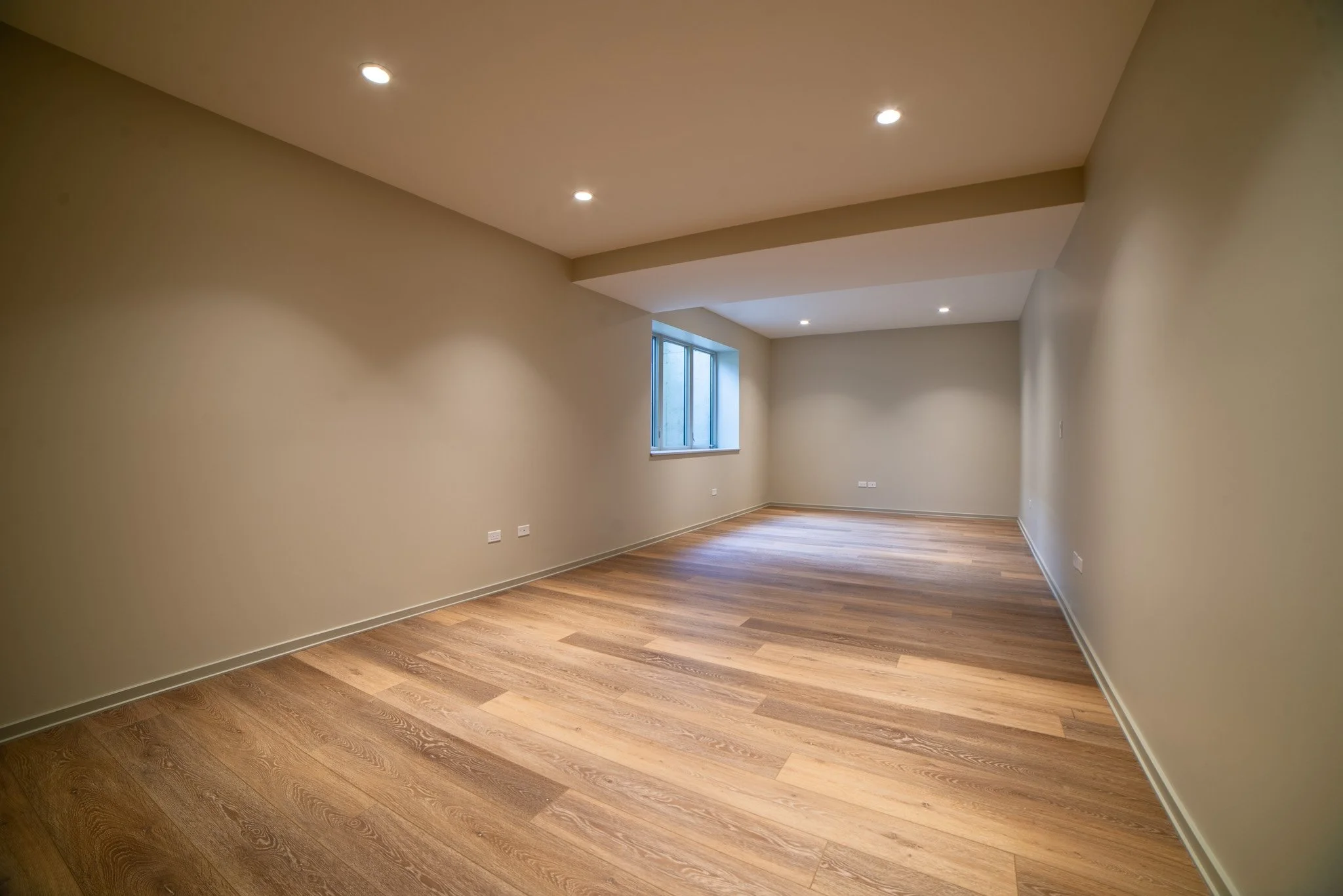 Empty room with wooden flooring, beige walls, small window, and ceiling recessed lights.