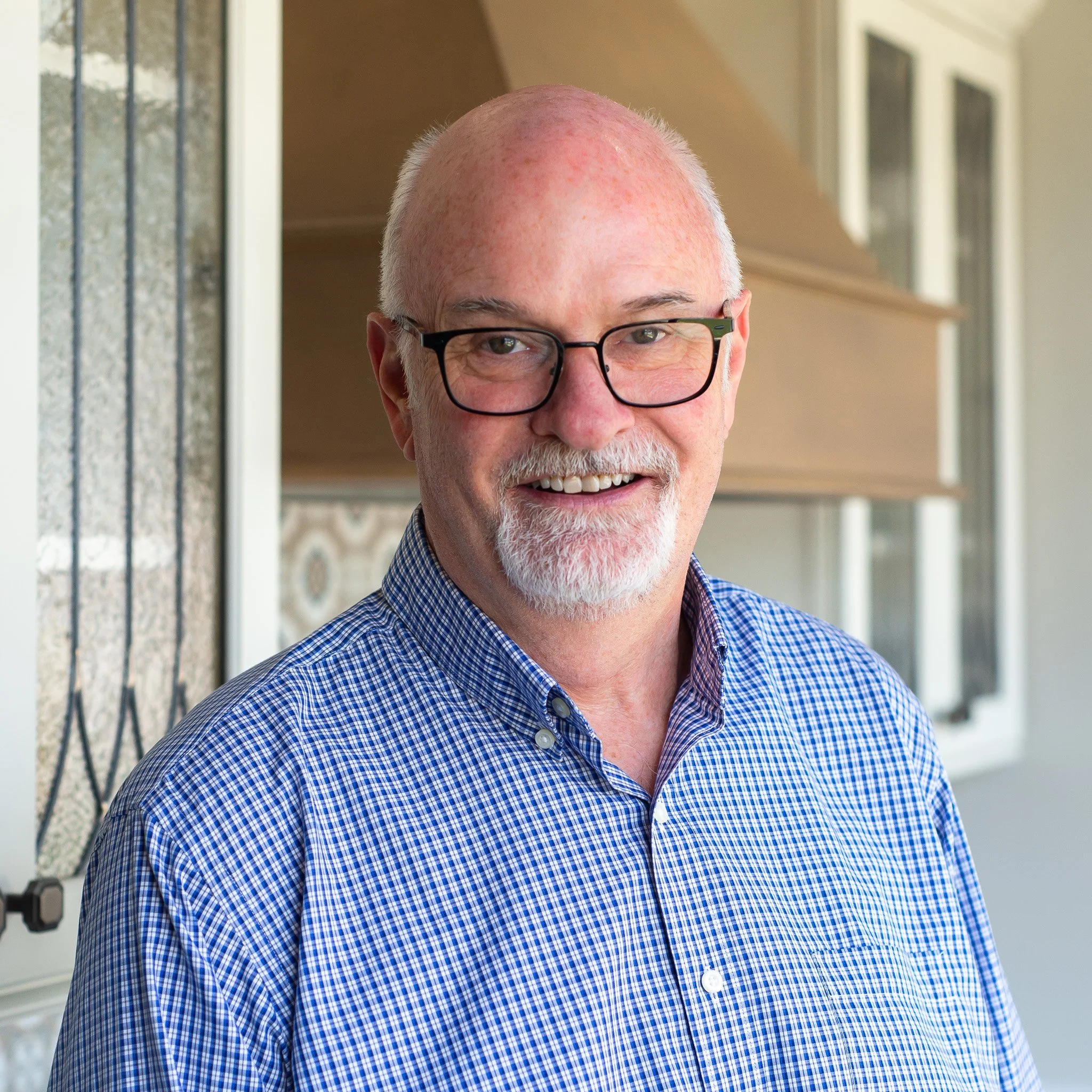 Close-up of smiling elderly man with glasses, a white beard, and a bald head, wearing a blue checkered shirt, standing outdoors near a building.
