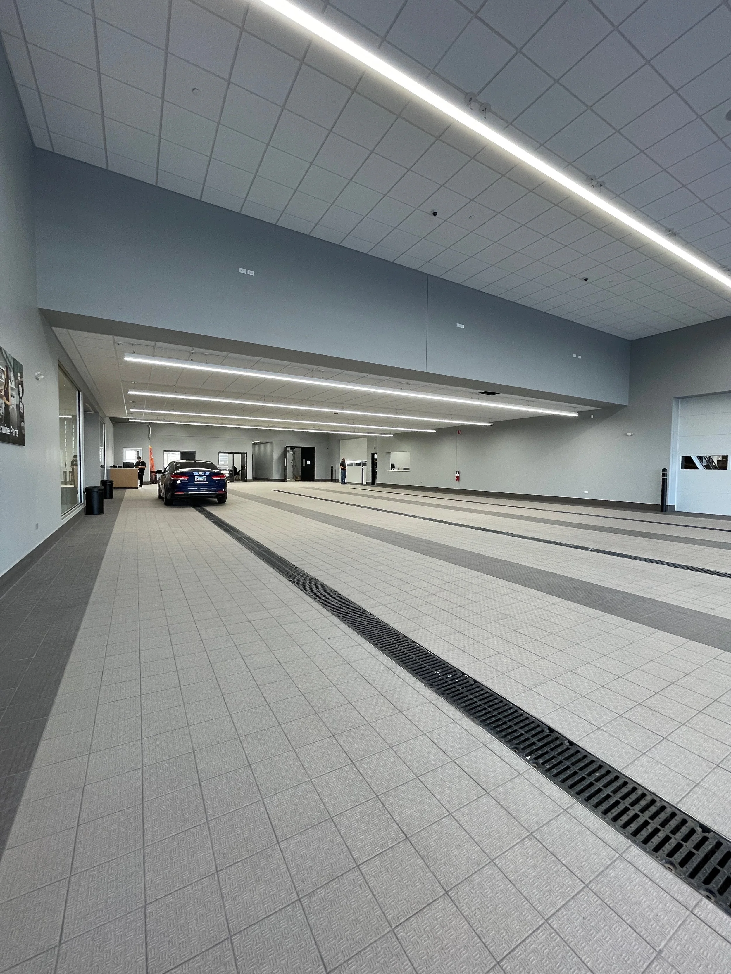 A large, clean indoor space resembling a garage or service area with tiled floors. A black car is parked near the wall, with a few people in the background. The ceiling has a suspended grid with linear lights, and there is a ventilation grate on the 