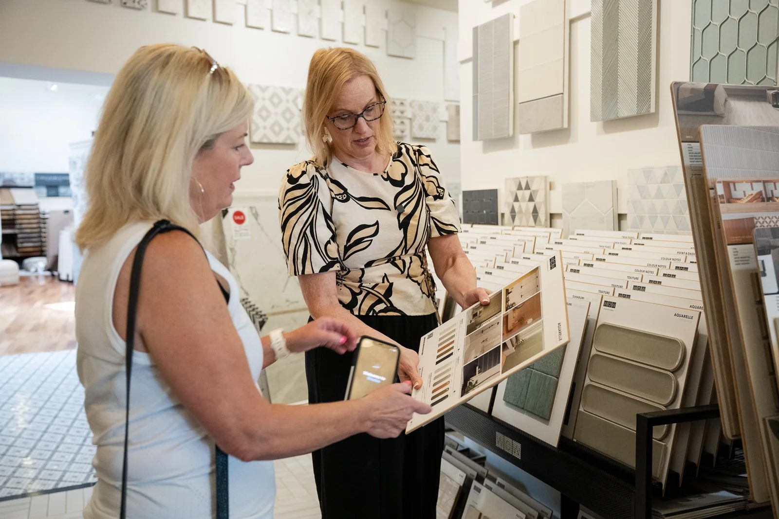 Two women shopping for tile samples in a home improvement store, with various ceramic and tile samples on display.