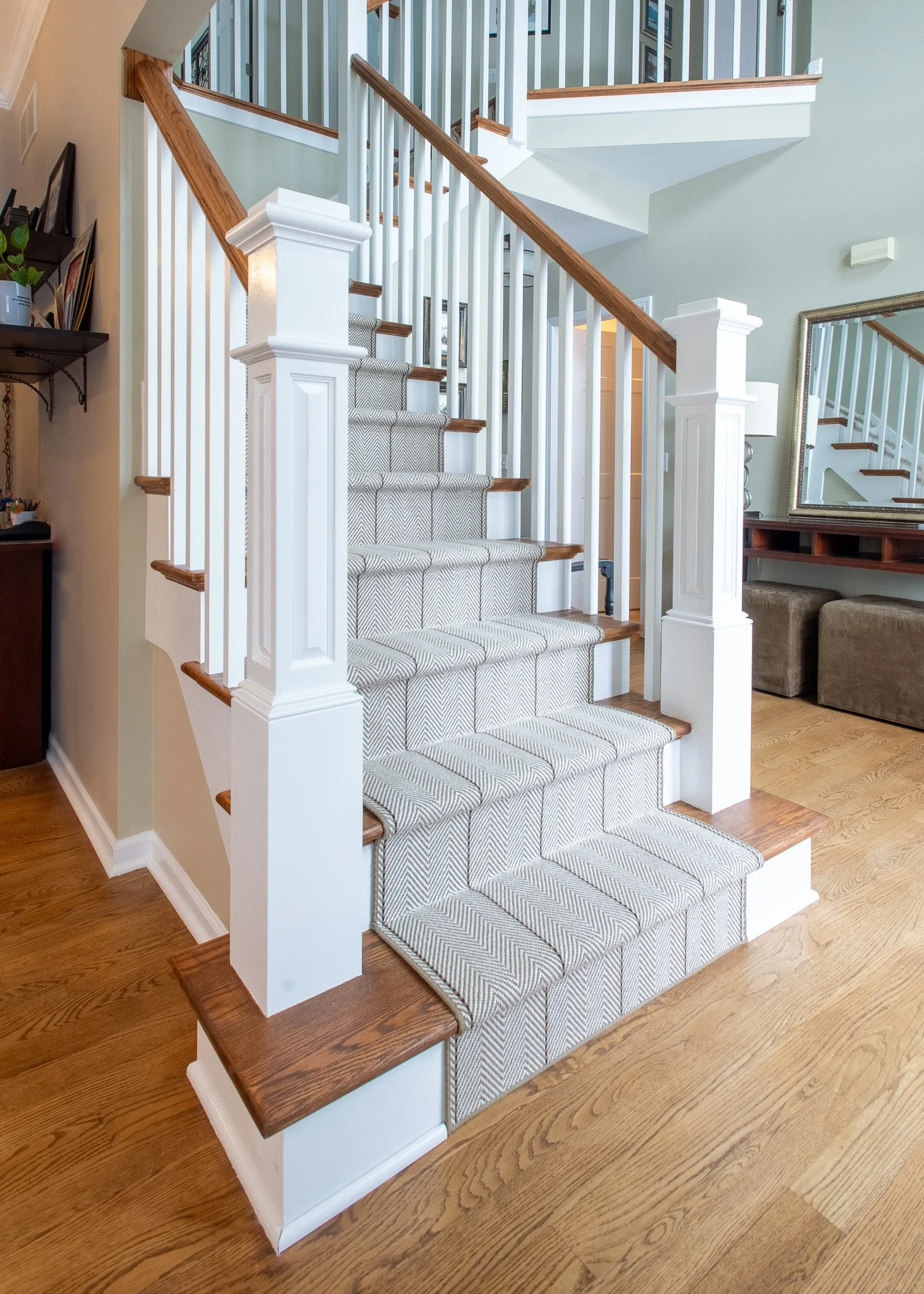 Interior view of a staircase with wooden steps and white spindles, a carpeted runner, wooden handrails, and a mirror reflecting part of the staircase, along with hardwood flooring.