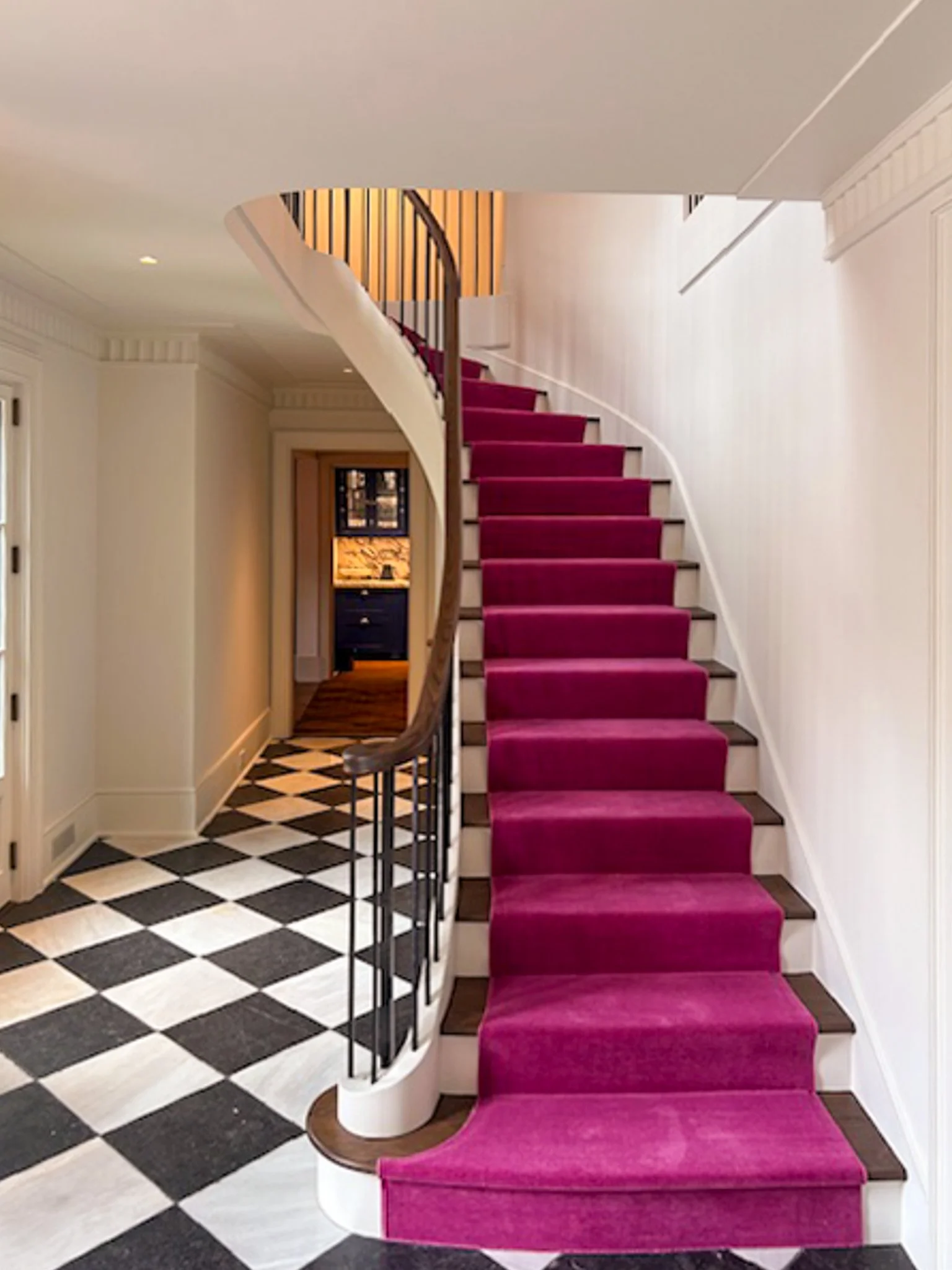 Interior of a house with a staircase with a plush pink carpet, white walls, black and white checkered flooring, and a corridor leading to another room.