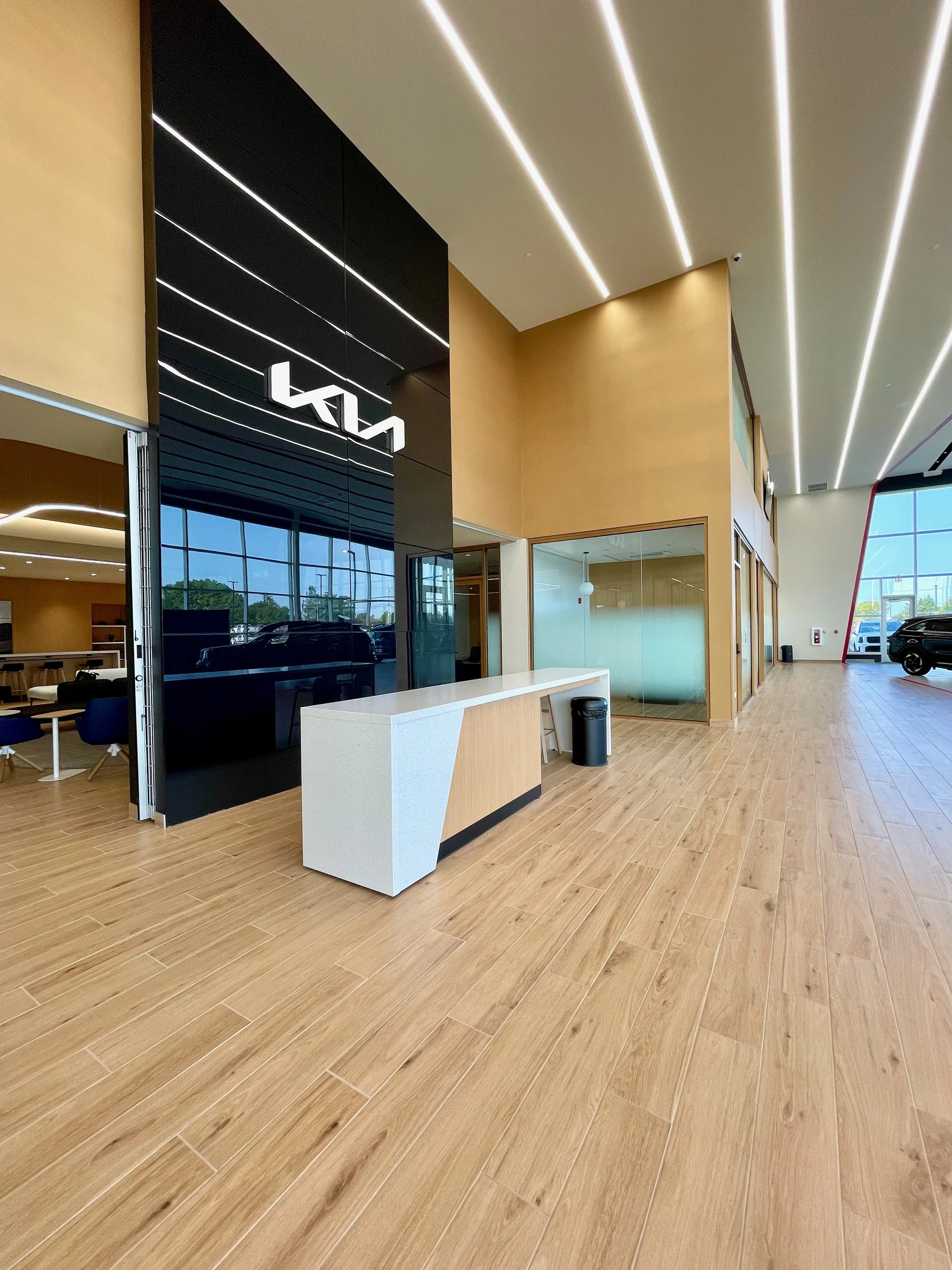 Interior of a car dealership with a modern reception desk, wooden flooring, large windows, and a black wall with a white logo.