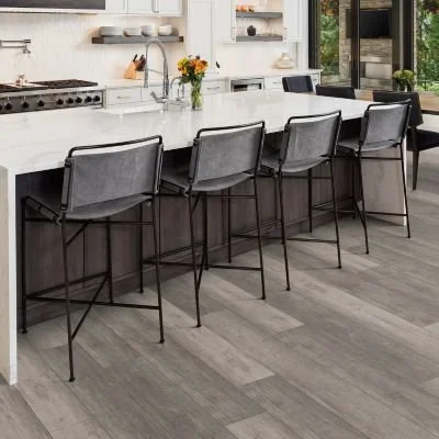 Modern kitchen with a white island and five gray bar stools, vase with flowers, and large windows showing greenery outside.
