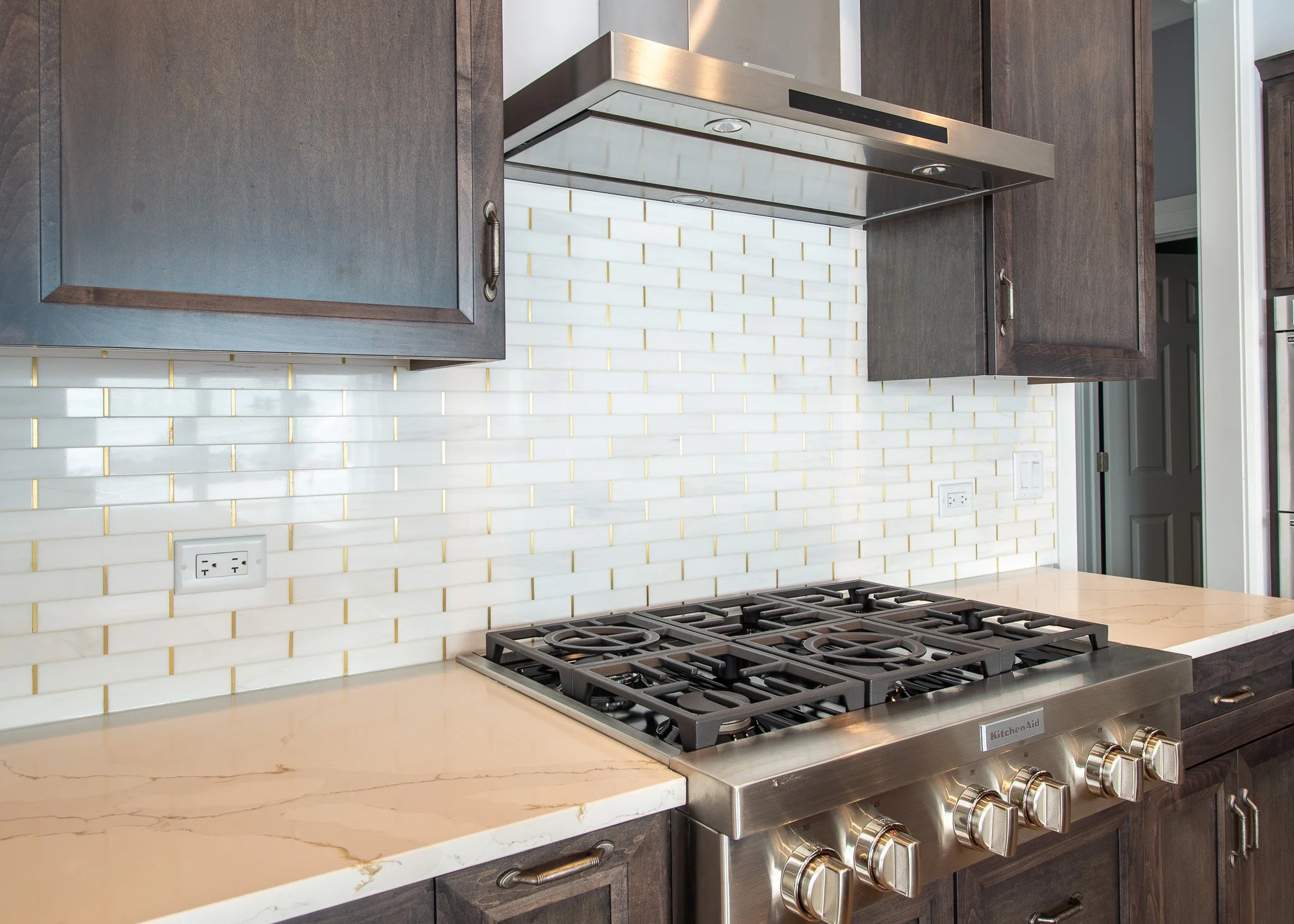 Modern kitchen with dark wood cabinets, white marble countertop, white tile backsplash with gold accents, stainless steel gas range and overhead vent hood.