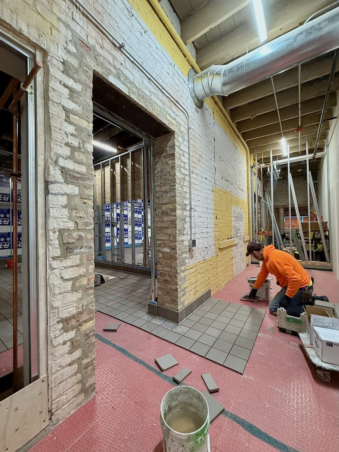 Construction worker installing gray tiles on the floor inside a building with exposed brick walls and ceiling pipes.
