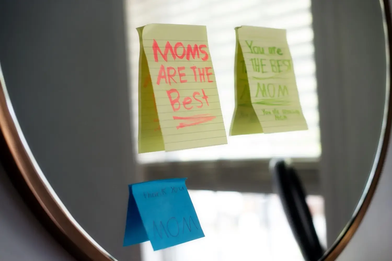 Colorful handwritten message notes on sticky notes attached to a mirror. The notes read: 'MOMS ARE THE BEST', 'You are the best MOM', and 'Thank you MOM'.