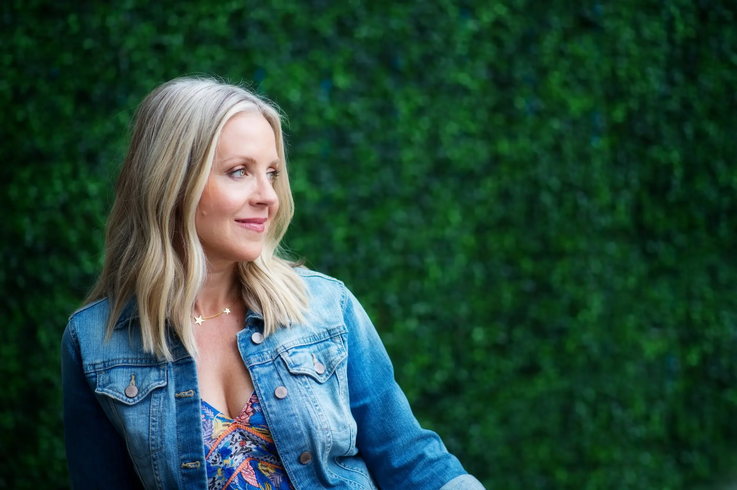 A woman with blonde hair, wearing a denim jacket and a colorful top, stands against a green leafy background, looking to her left with a slight smile.