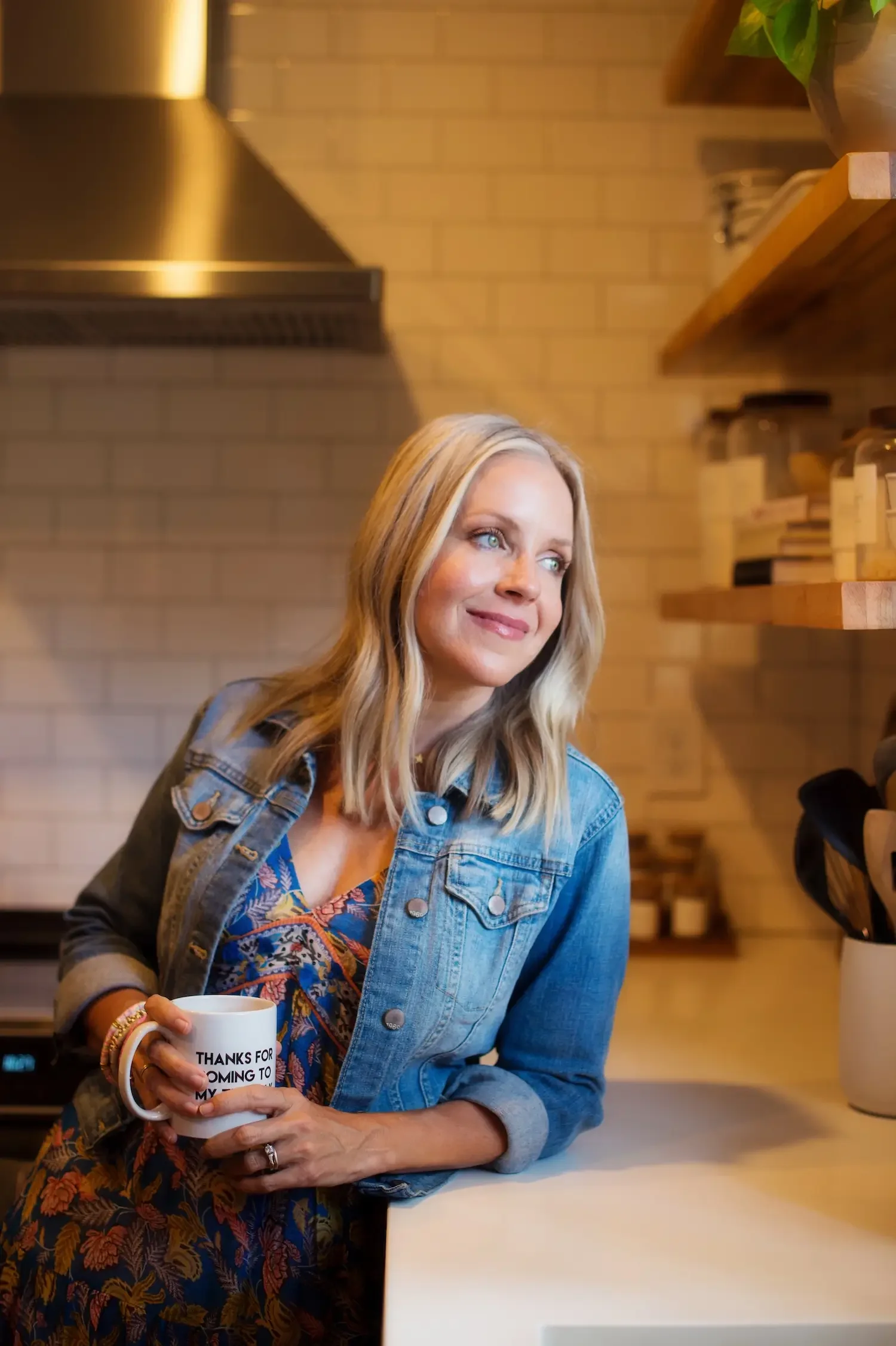 Angie Mizzell sitting at a white table in a cozy kitchen, holding a white mug that says 'Thanks for coming to my Ted Talk'.