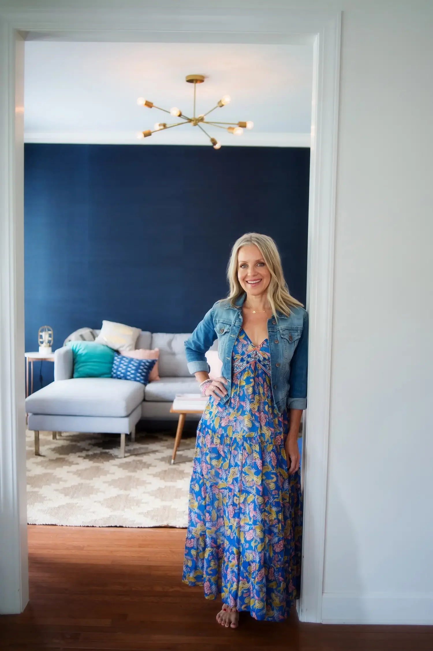 Angie Mizzell standing in the doorway to her home office, wearing a blue and yellow floral maxi dress and denim jacket, smiling at the camera.