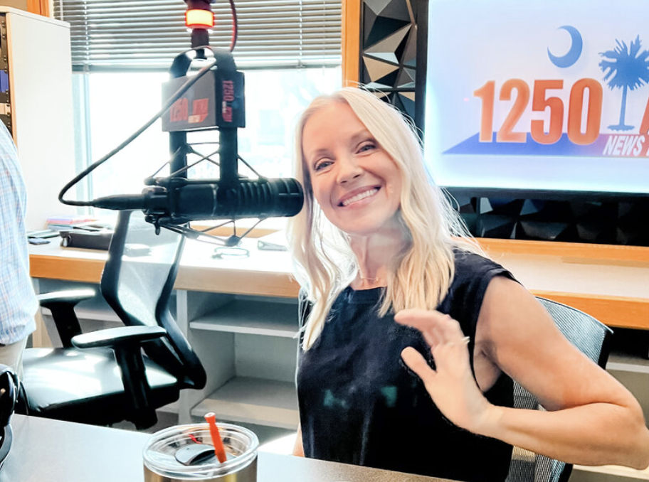 Angie Mizzell is waving in a radio studio with a microphone and a screen displaying '1250 News' in the background.