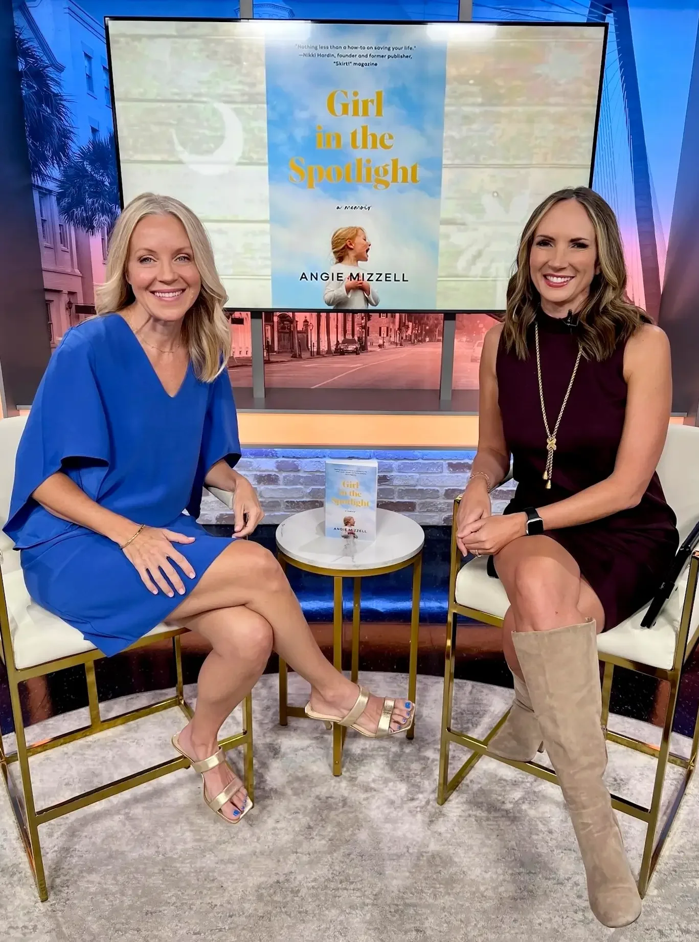  Angie Mizzell on a talk show set with another woman, both smiling at the camera. Behind them is a large screen displaying a book cover titled 'Girl in the Spotlight' by Angie Mizzell.
