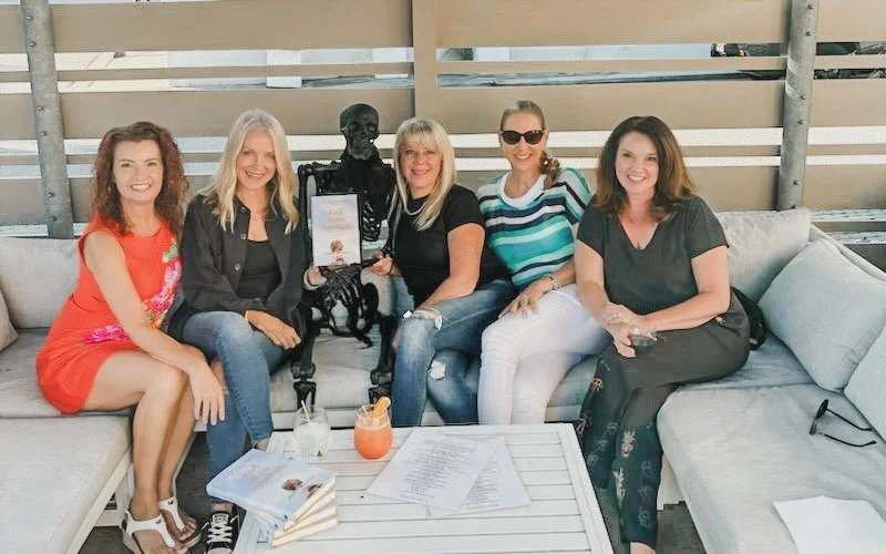 Angie Mizzell at a book club meeting with four other women, sitting on a patio sofa with a skeleton prop, Angie's books 'Girl in the Spotlight', and drinks in front of them.