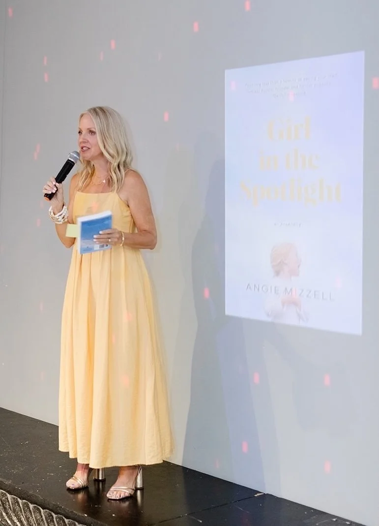 Woman in a yellow dress holding a microphone and a booklet, standing on a stage with a screen displaying a book titled "Girl in the Spotlight" by Angie Mizzell.