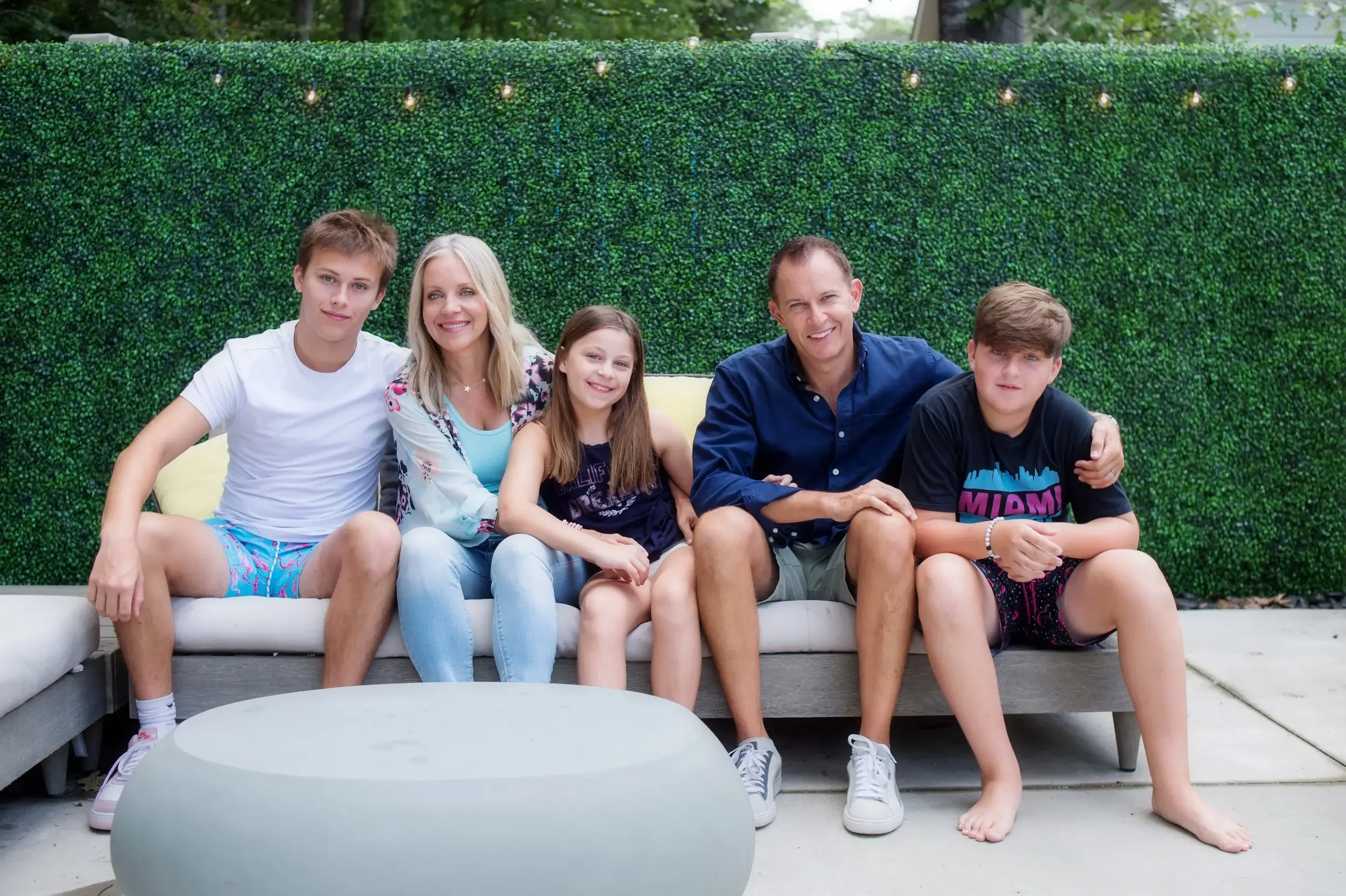 A family of six sitting together on outdoor sofa, smiling, against a green hedge background.