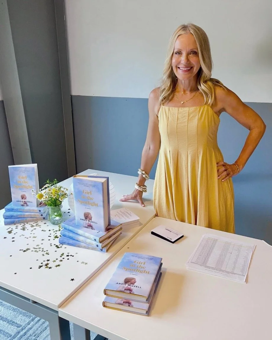 Angie Mizzell stands behind a table with stacks of her book titled 'Girl in the Spotlight', a small bouquet of yellow and white flowers, and some gold star-shaped confetti. She is smiling and has long blonde hair.