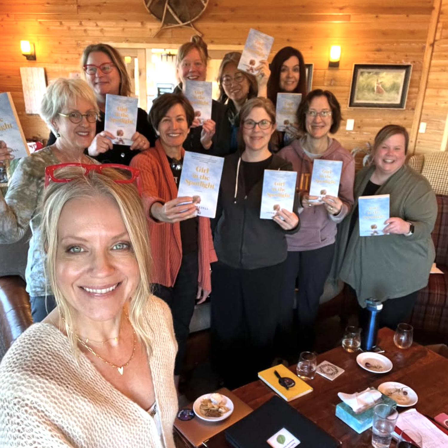 A group of women gathered in a cozy wooden room, holding copies of a book titled 'Girl in the Spotlight'. The woman in the front is Angie Mizzell, author of the book.