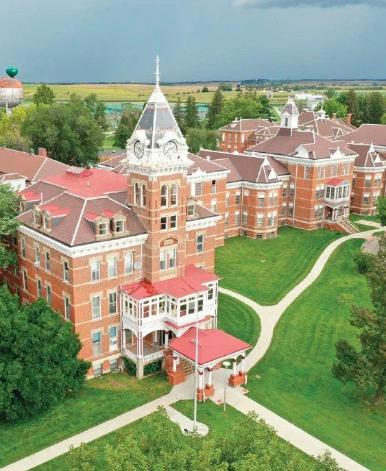 A large historic brick hospital with a tower and a wraparound porch, surrounded by green lawns and walking paths.